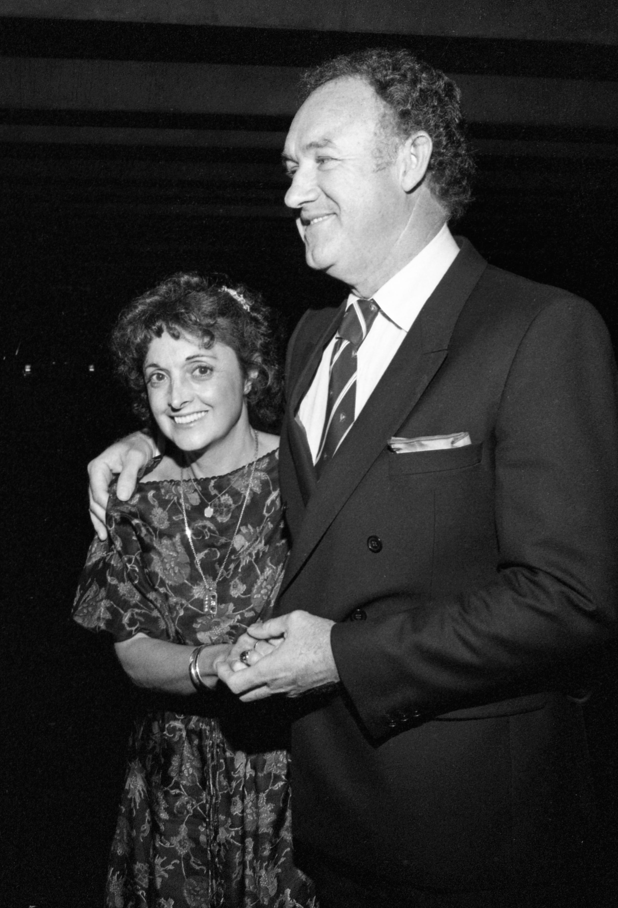 Faye Maltese and Gene Hackman pose together at an event while holding hands. Maltese wears a floral dress, and Hackman wears a suit and tie
