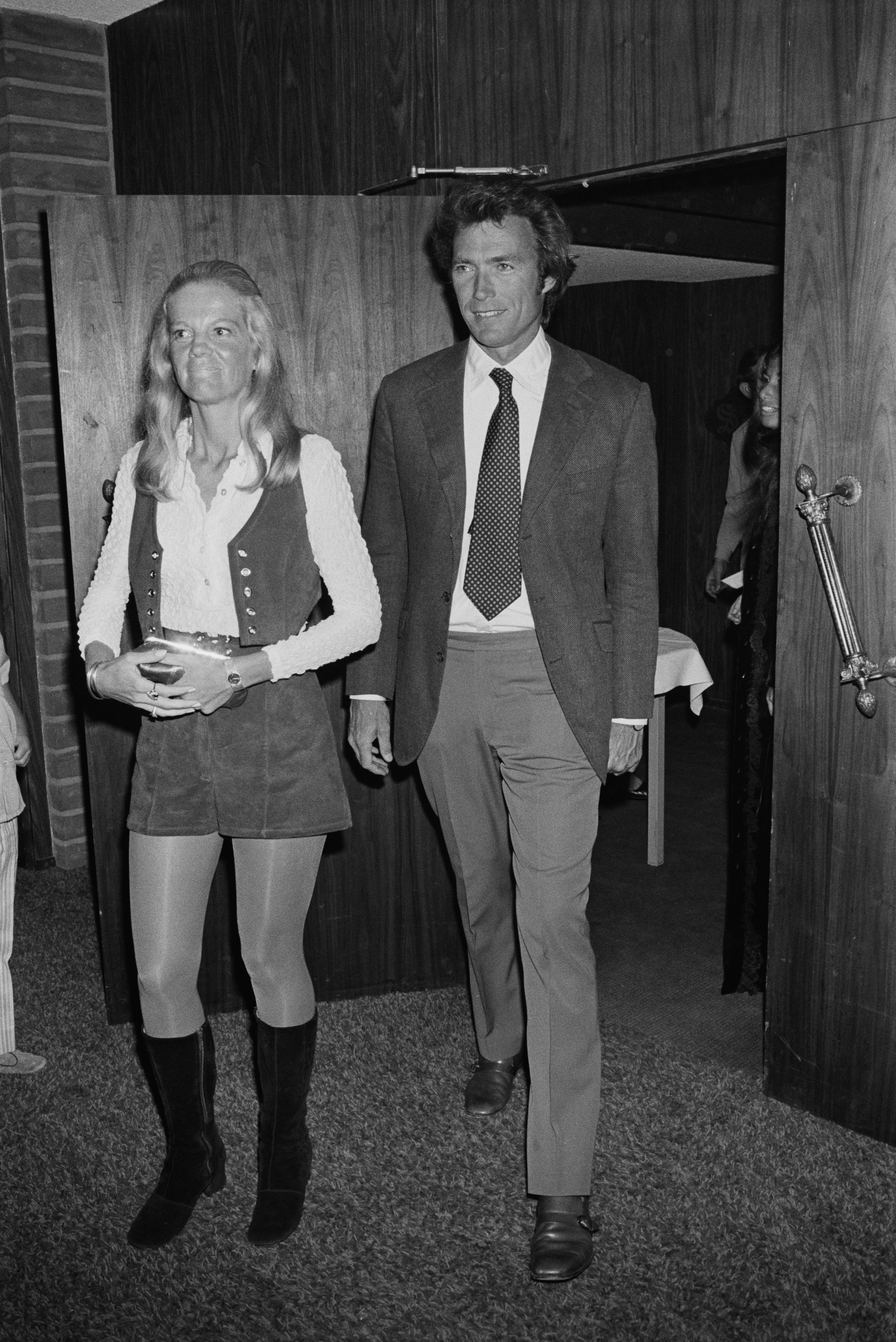 Maggie Johnson wears a stylish vest and skirt with knee-high boots and walks beside Clint Eastwood, who wears a suit and tie, exiting a wooden double door