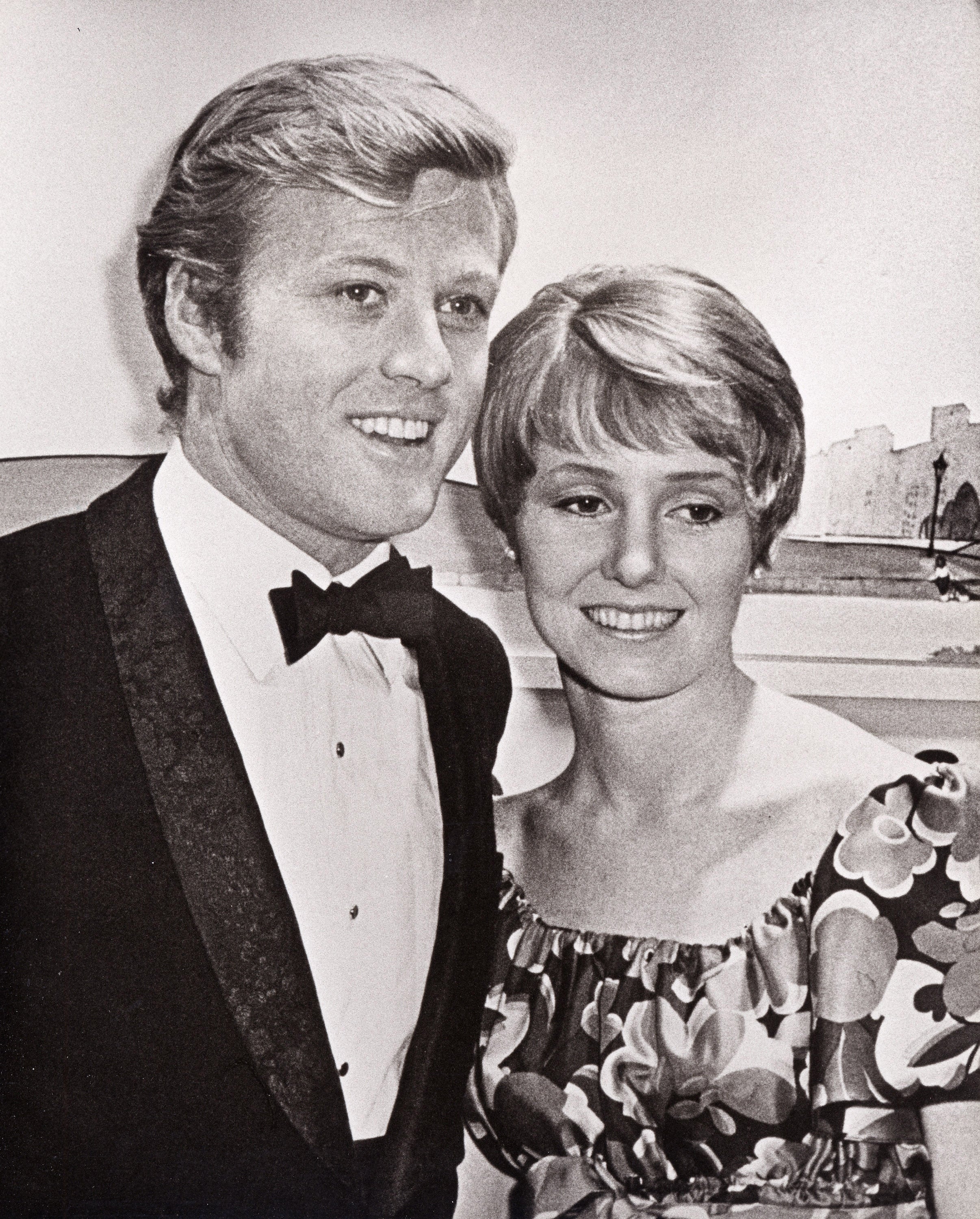 Robert Redford and Lola Van Wagenen pose together. Redford wears a tuxedo, and Van Wagenen wears a floral dress