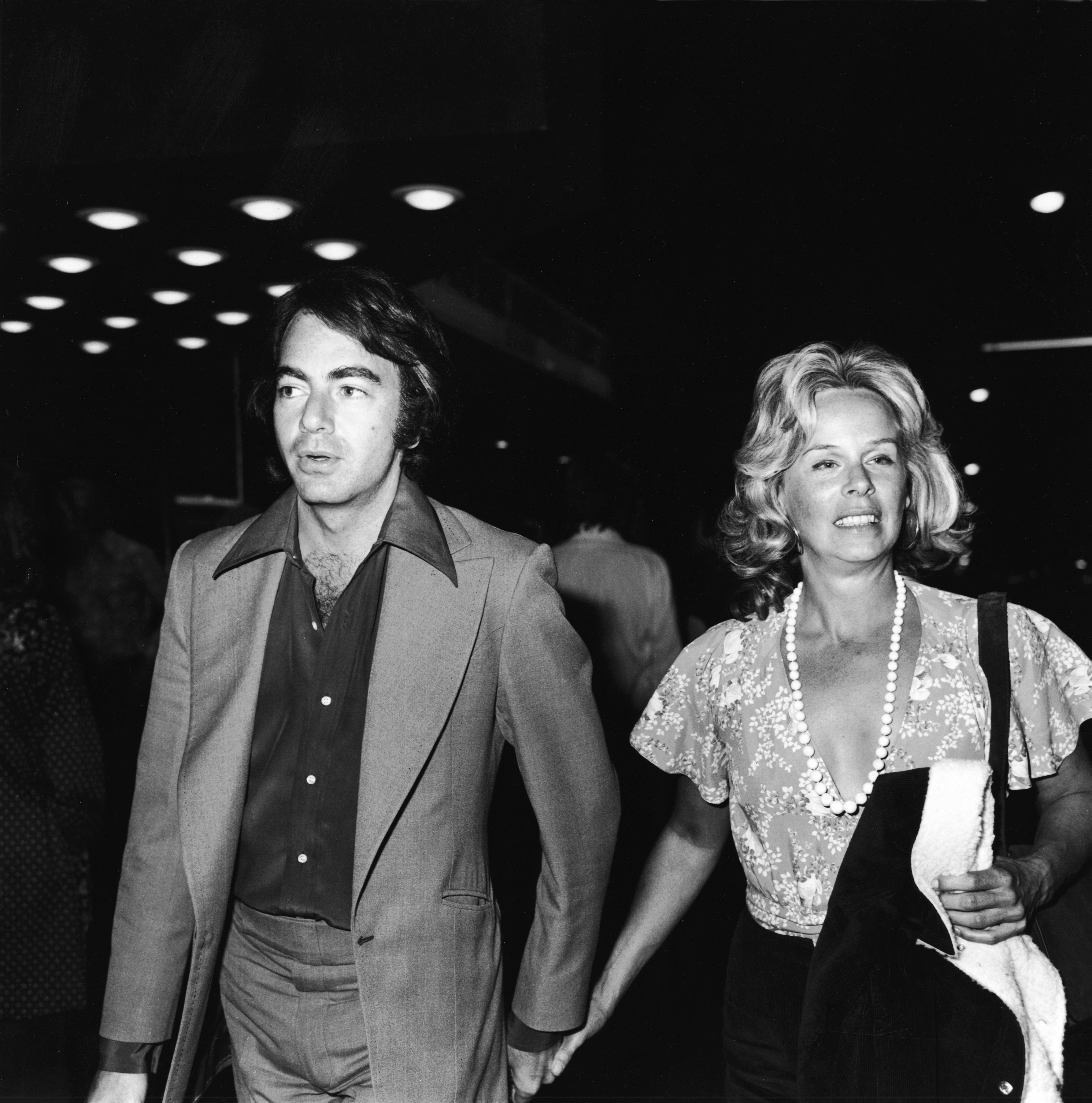 Neil Diamond and Marcia Murphey hold hands while walking at an event. Diamond wears a suit with a bold collar and Murphey wears a blouse with a large string of pearls