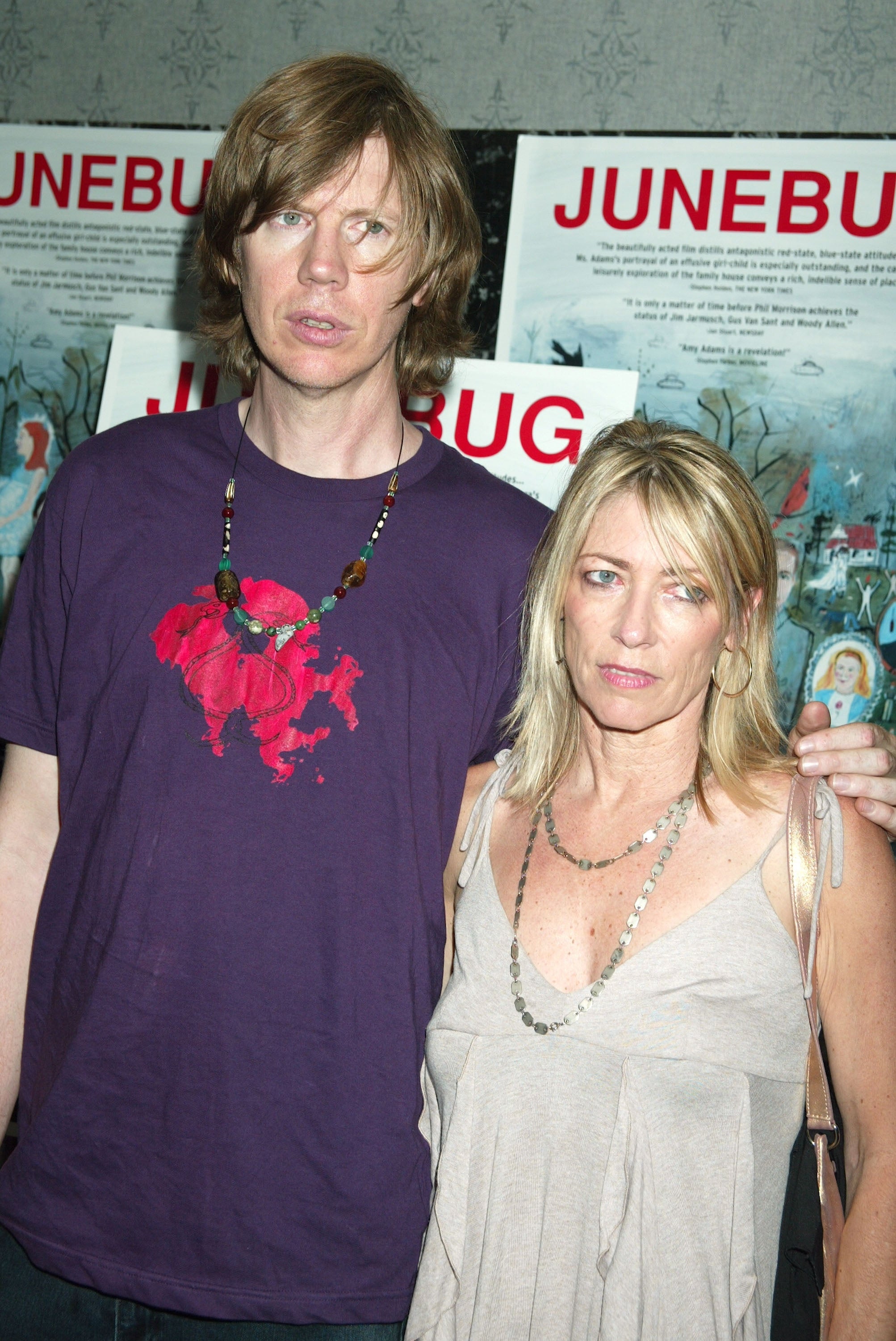 Thurston Moore and Kim Gordon stand in front of "Junebug" posters. Moore wears a graphic tee, and Gordon wears a sleeveless dress