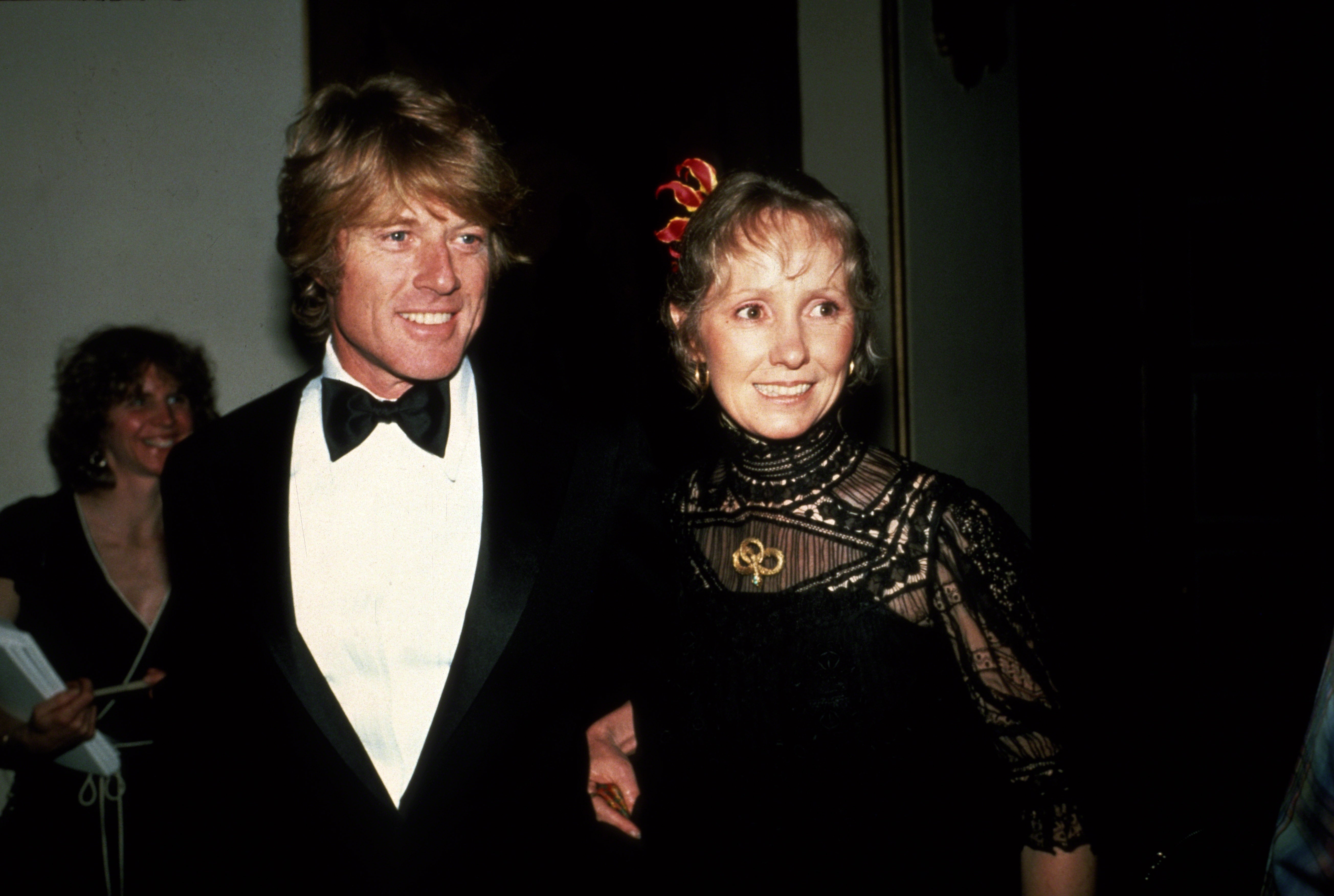 Robert Redford and Lola Van Wagenen pose together at a formal event. Redford wears a classic tuxedo, and Van Wagenen wears an elegant lace dress