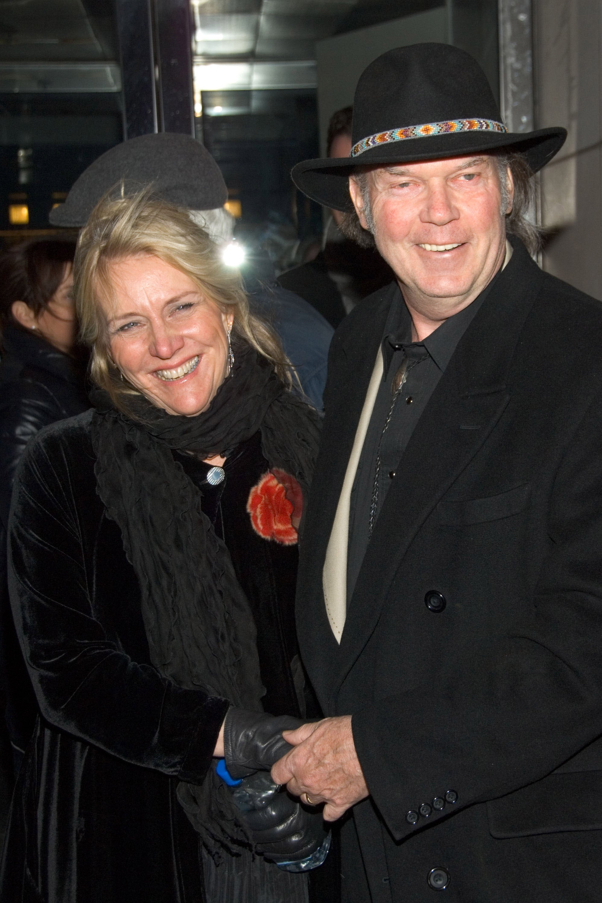 Pegi Young and Neil Young smile while holding hands. Neil wears a dark coat and a wide-brimmed hat. Pegi wears a coat with a flower pin