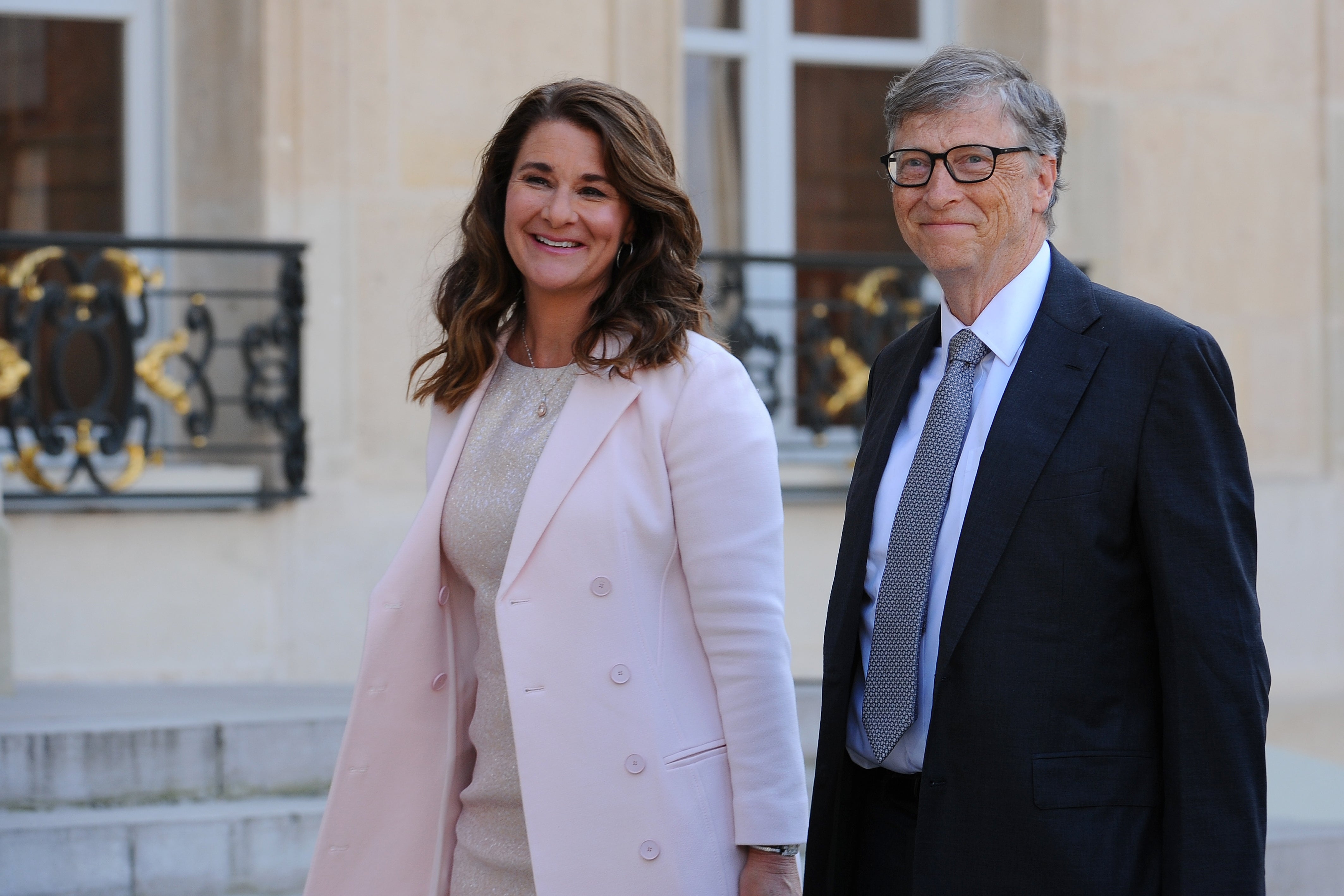 Melinda French Gates and Bill Gates walk together near an ornate building. French Gates wears a light pink trench coat and a tan dress. Gates wears a suit with a grey tie