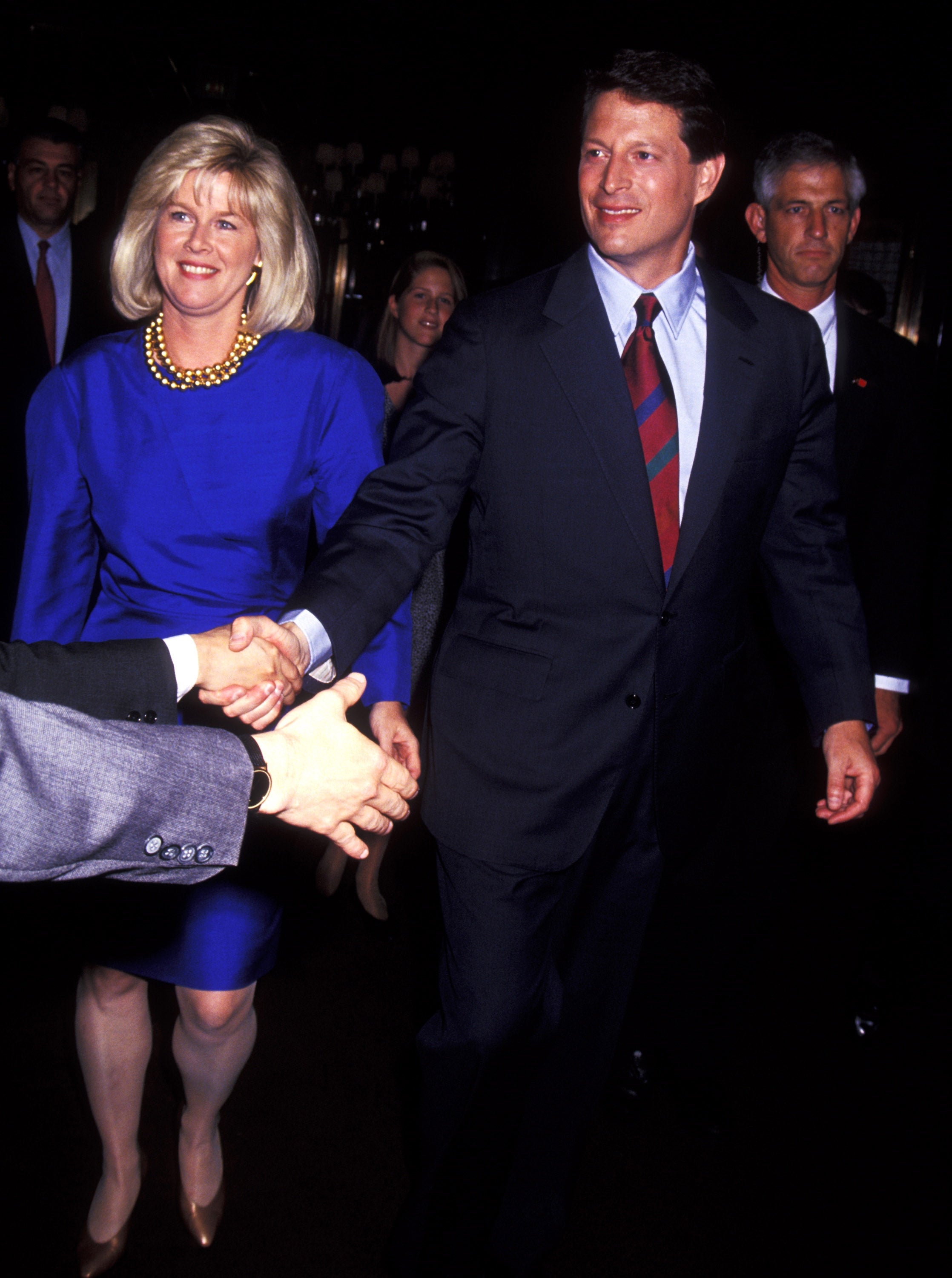 Al Gore wears a formal suit and shakes hands at an event. Tipper Gore walks beside him and wears a stylish, knee-length dress with a pearl necklace