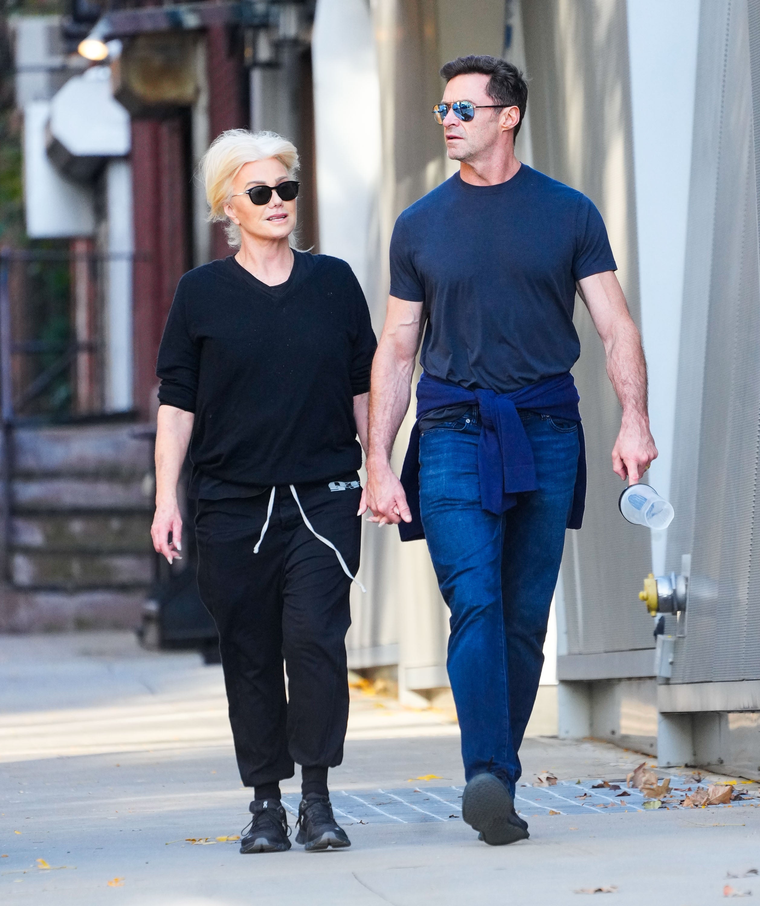 Deborra-lee Furness and Hugh Jackman walk hand in hand on a sidewalk, both wearing casual outfits with sunglasses; Furness in a black top and pants, and Jackman in a dark shirt with jeans