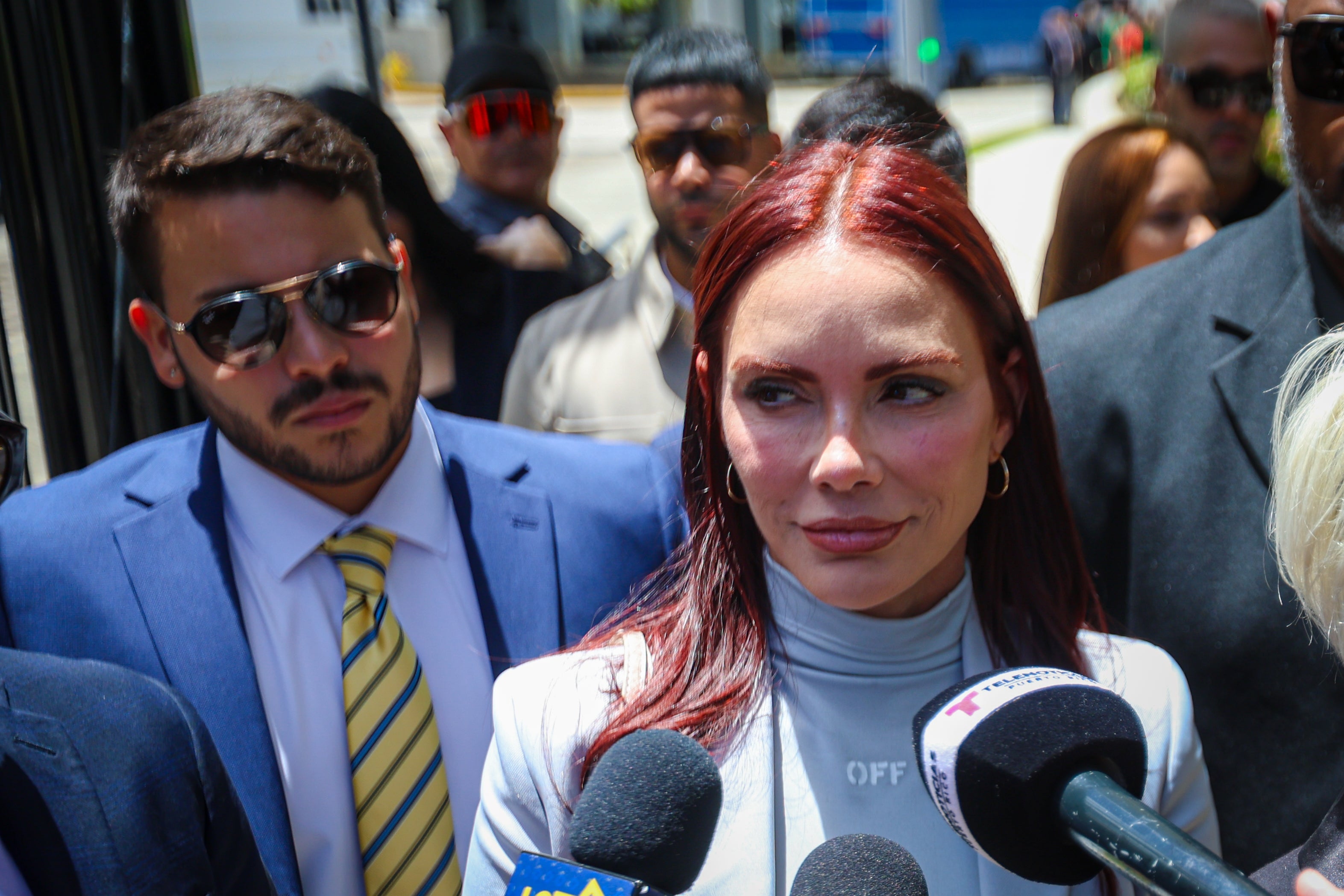 Mireddys González wears a stylish coat and stands confidently behind multiple microphones, surrounded by suited individuals outdoors