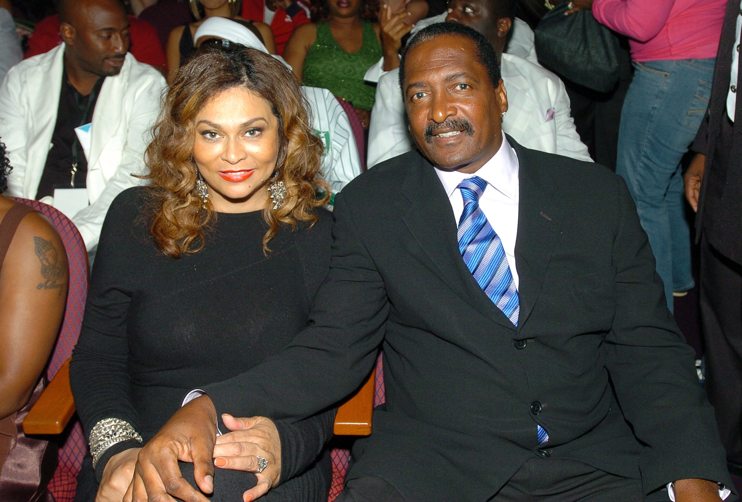 Tina Knowles and Matthew Knowles sit together at an event, holding hands. Tina wears a black outfit, and Matthew wears a suit with a striped tie. Other attendees are in the background