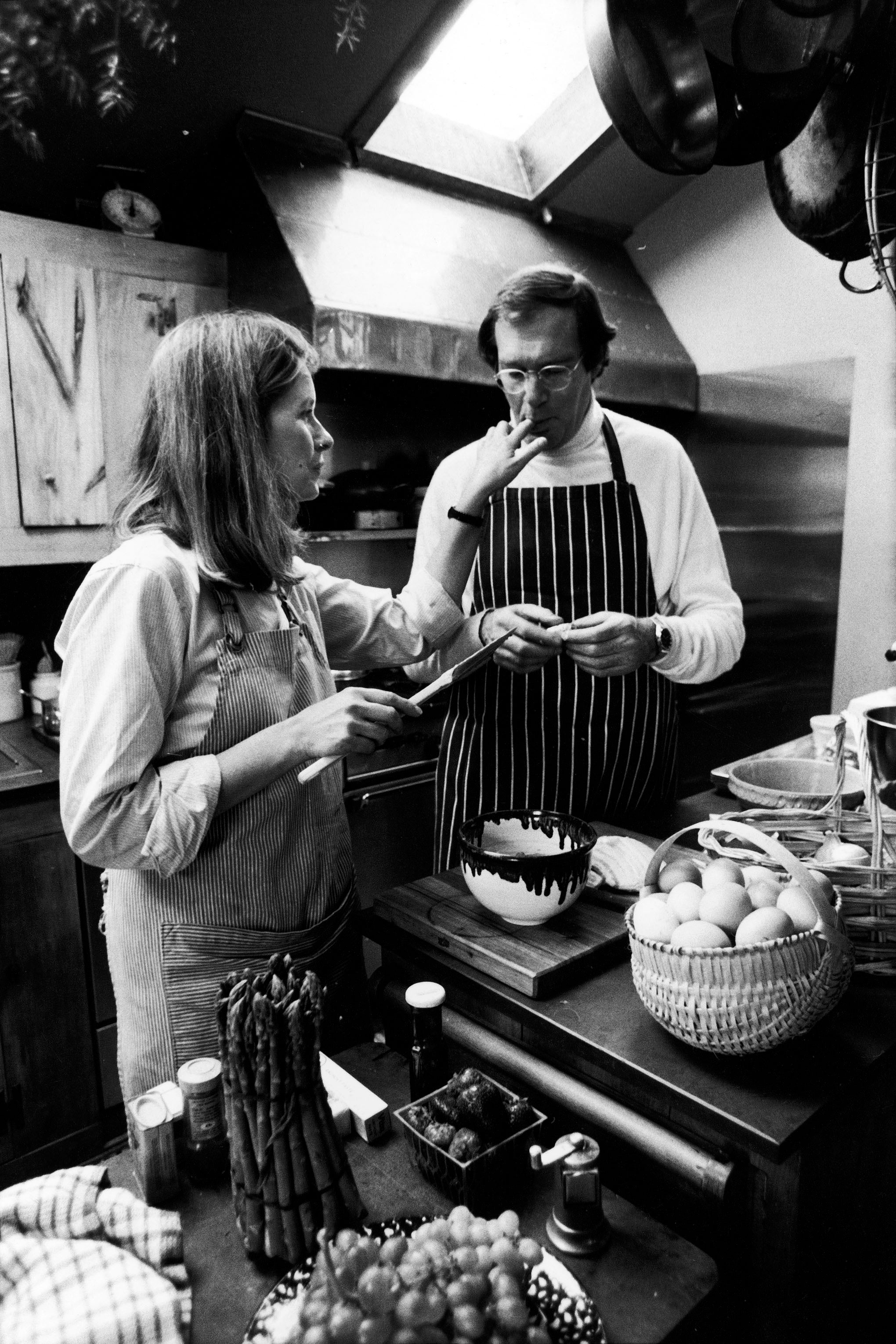 Martha Stewart and Andrew Stewart wear aprons in a kitchen, preparing food together. The counter is filled with fresh produce and kitchen utensils