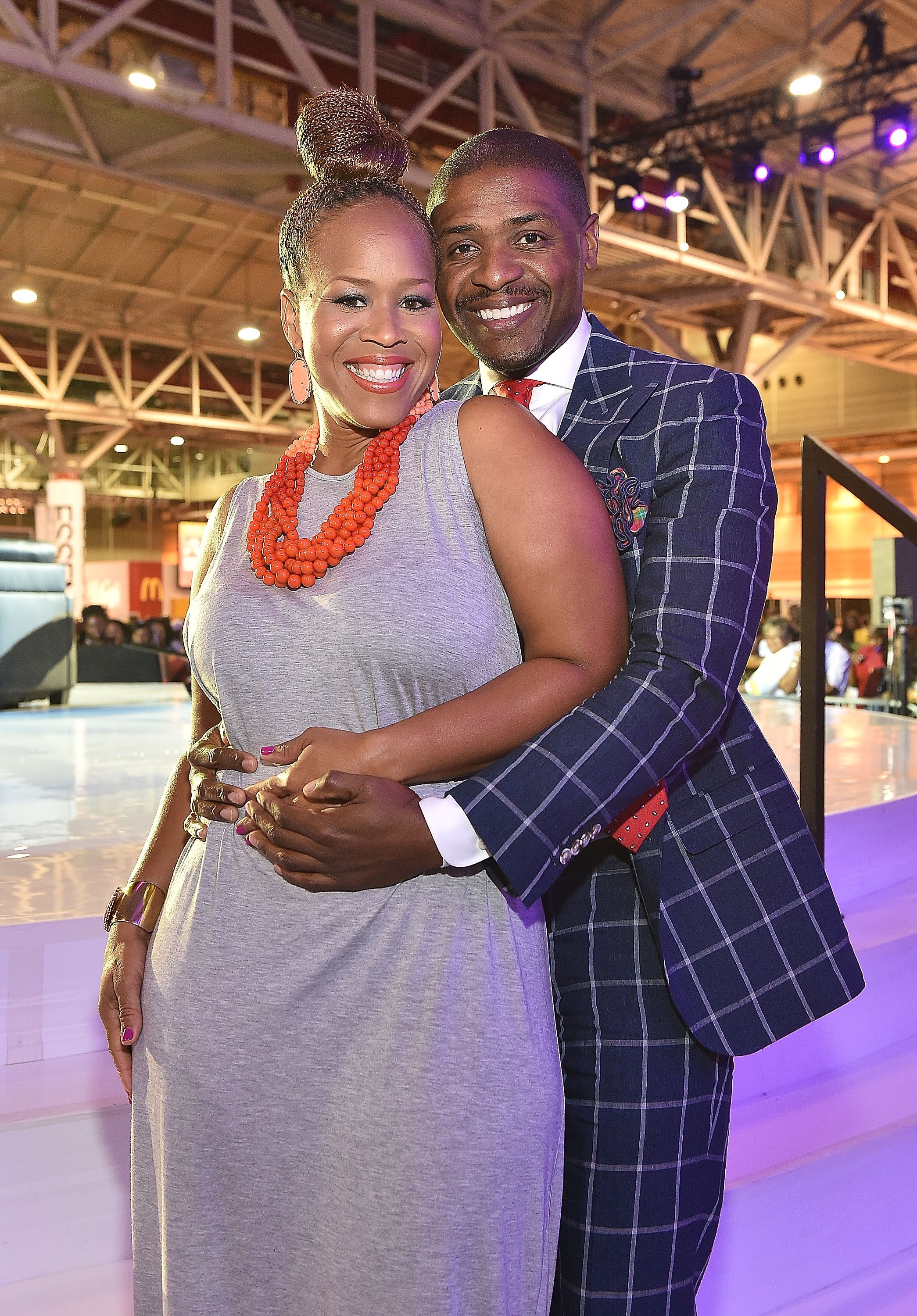 Tina and Teddy Campbell smile while embracing at an indoor event. Tina wears a gray dress with a large statement necklace, and Teddy wears a checkered suit