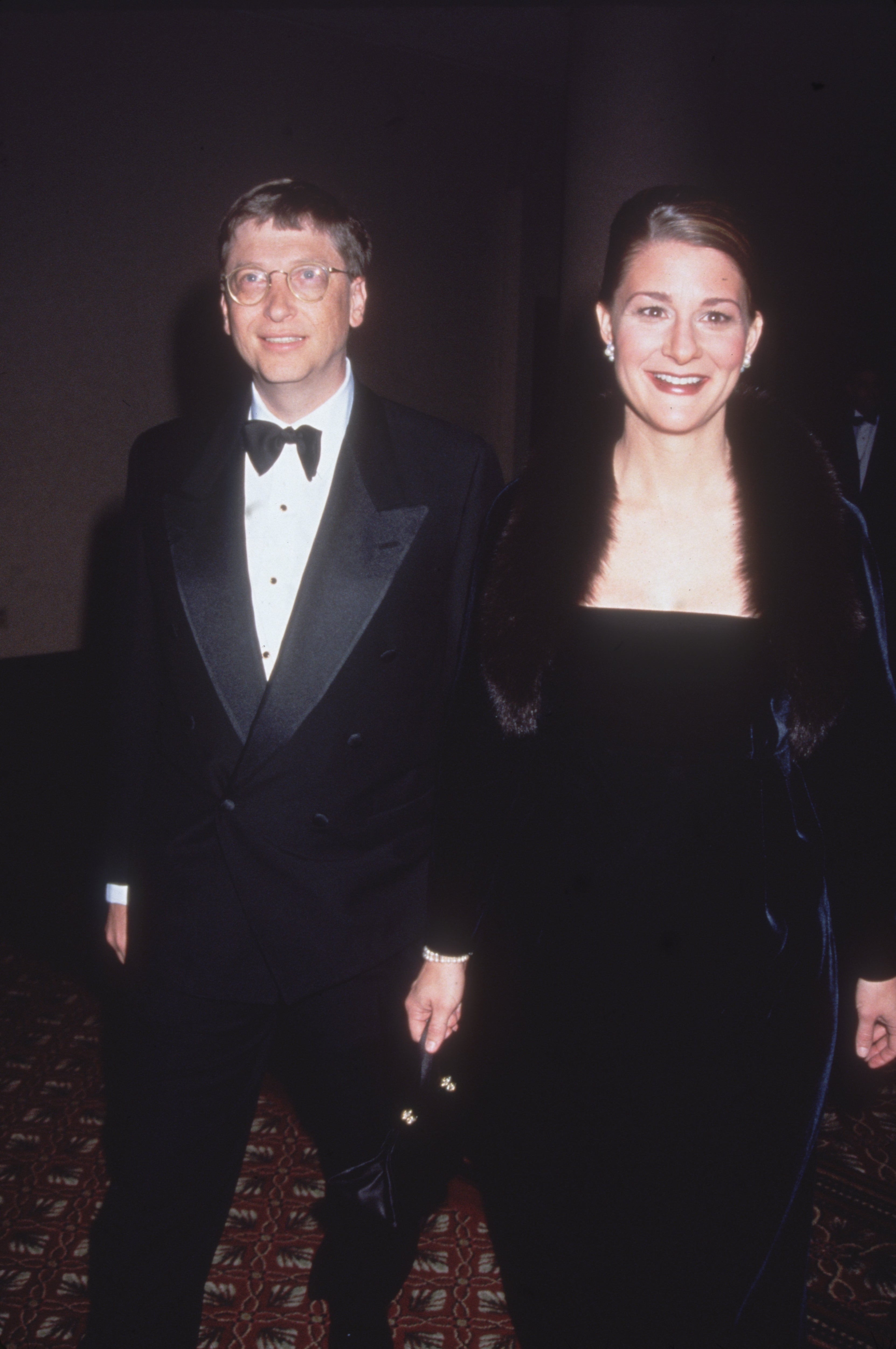 Bill Gates and Melinda French Gates walk together in formal evening attire. Gates wears a tuxedo, and French Gates wears a gown a fur shawl