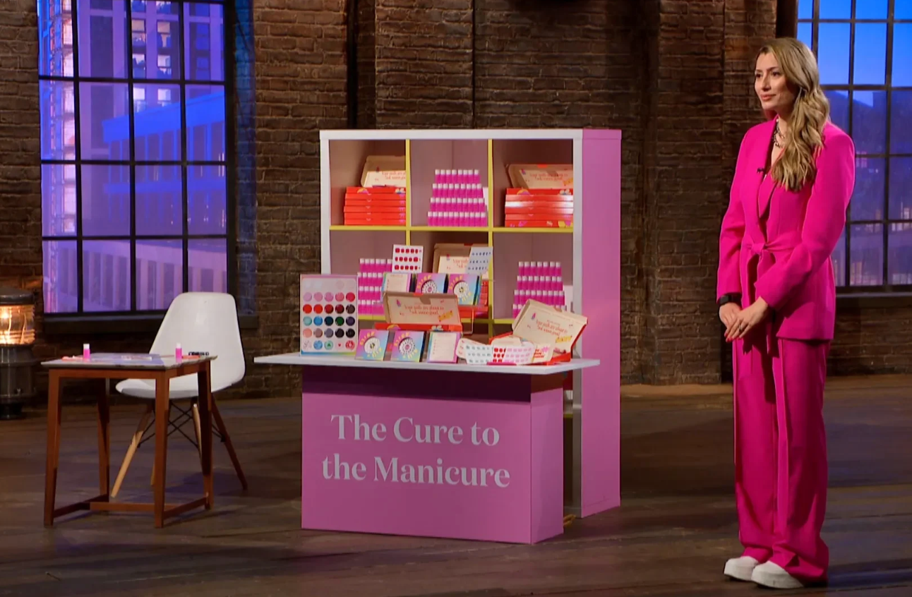 En person i en stilig rosa buksedress står ved siden av en skjerm med manikyrprodukter på et talkshow-sett