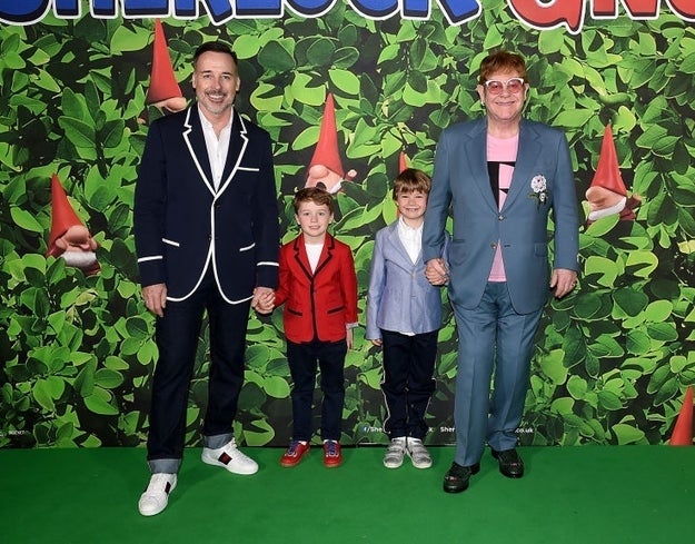 Four people stand in front of a gnome-themed backdrop, including individuals dressed in casual and formal attire holding hands