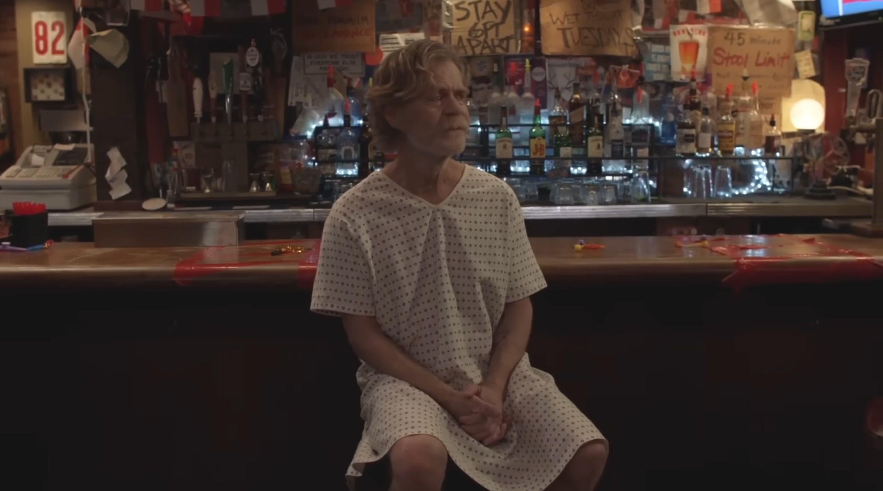Person in a hospital gown sitting at a bar, surrounded by various bottles and decorations