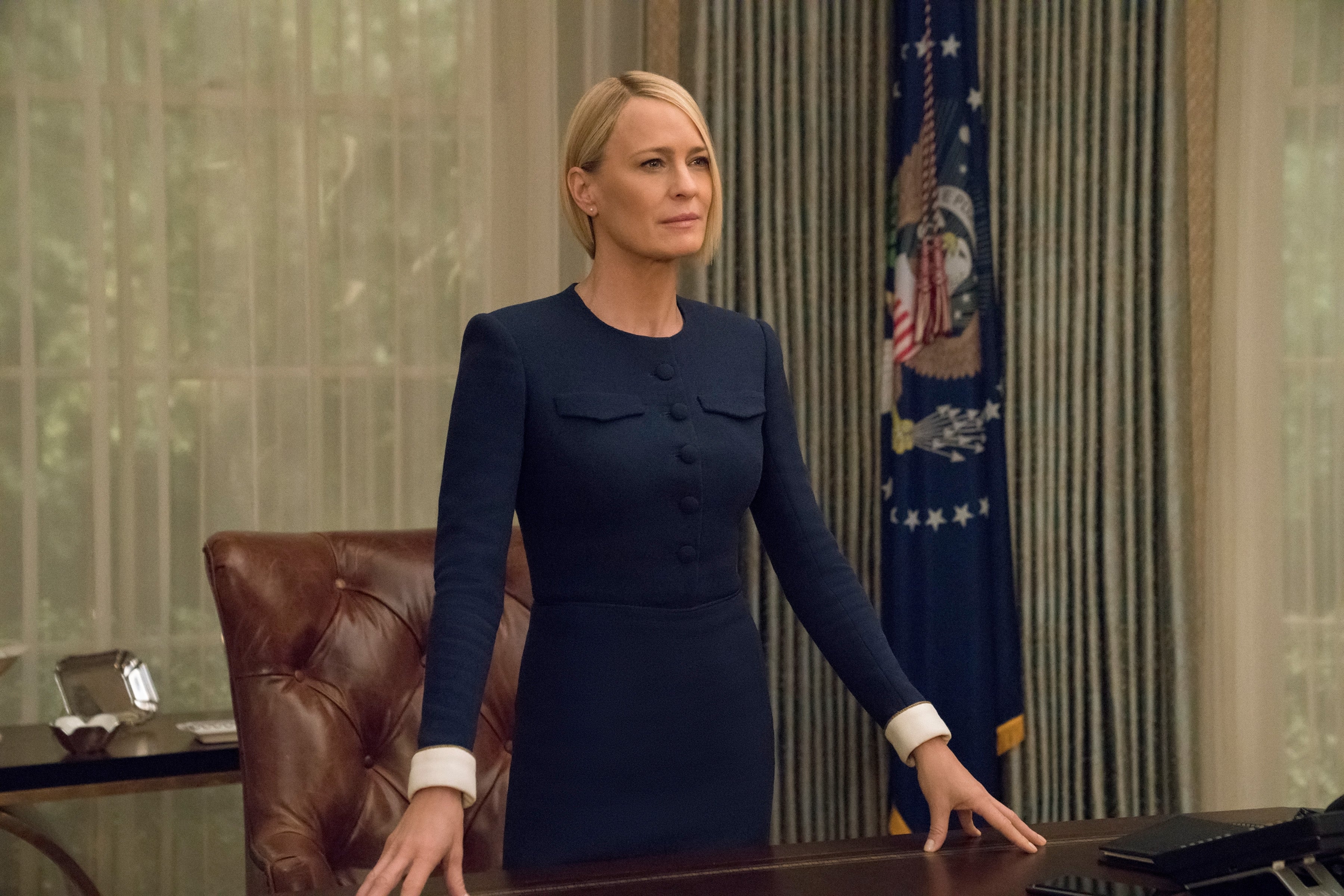 Person in a formal setting, wearing a tailored dark suit, standing authoritatively at a desk with a flag and curtains in the background