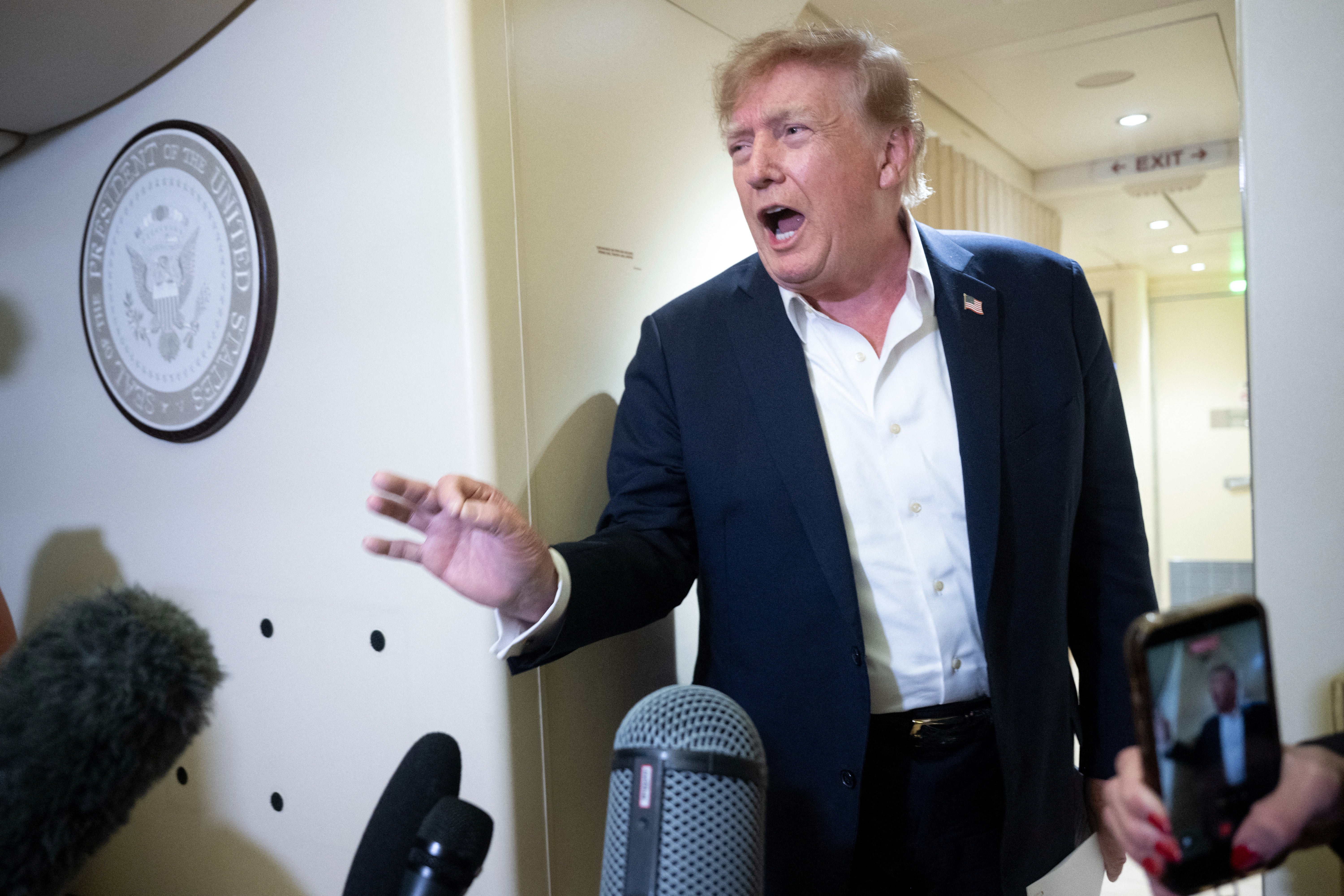 En person i dress snakker lidenskapelig til journalister på et fly, omgitt av mikrofoner og kameraer. Presidentsegl synlig på veggen