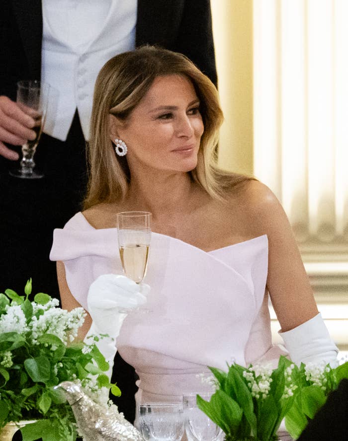 A woman at an elegant event holding a champagne glass, wearing an off-shoulder gown, white gloves, and sparkling earrings, surrounded by greenery