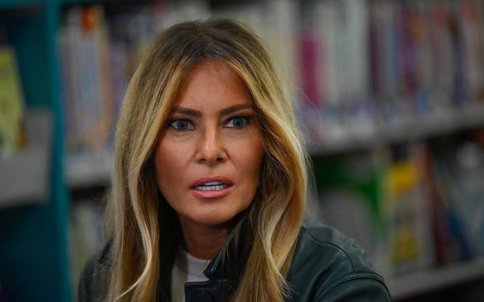 A woman with long hair sits in front of blurred bookshelves, wearing a leather jacket, looking engaged