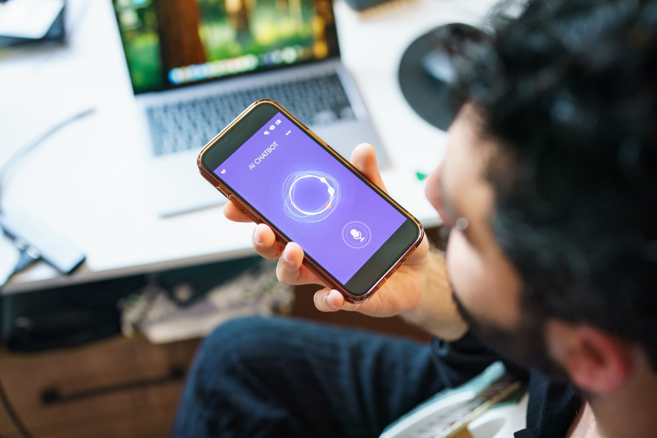 Mann som bruker en ai chatbot-app på en smarttelefon med en bærbar datamaskin synlig i bakgrunnen