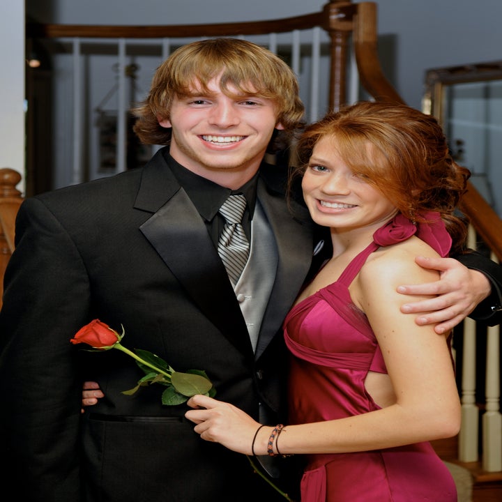 Two people smiling; one in a suit holding a rose, the other in an elegant gown with a halter neckline, posing together