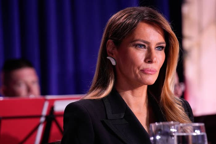 A person in formal attire is seated at an event with a focused expression, wearing statement earrings