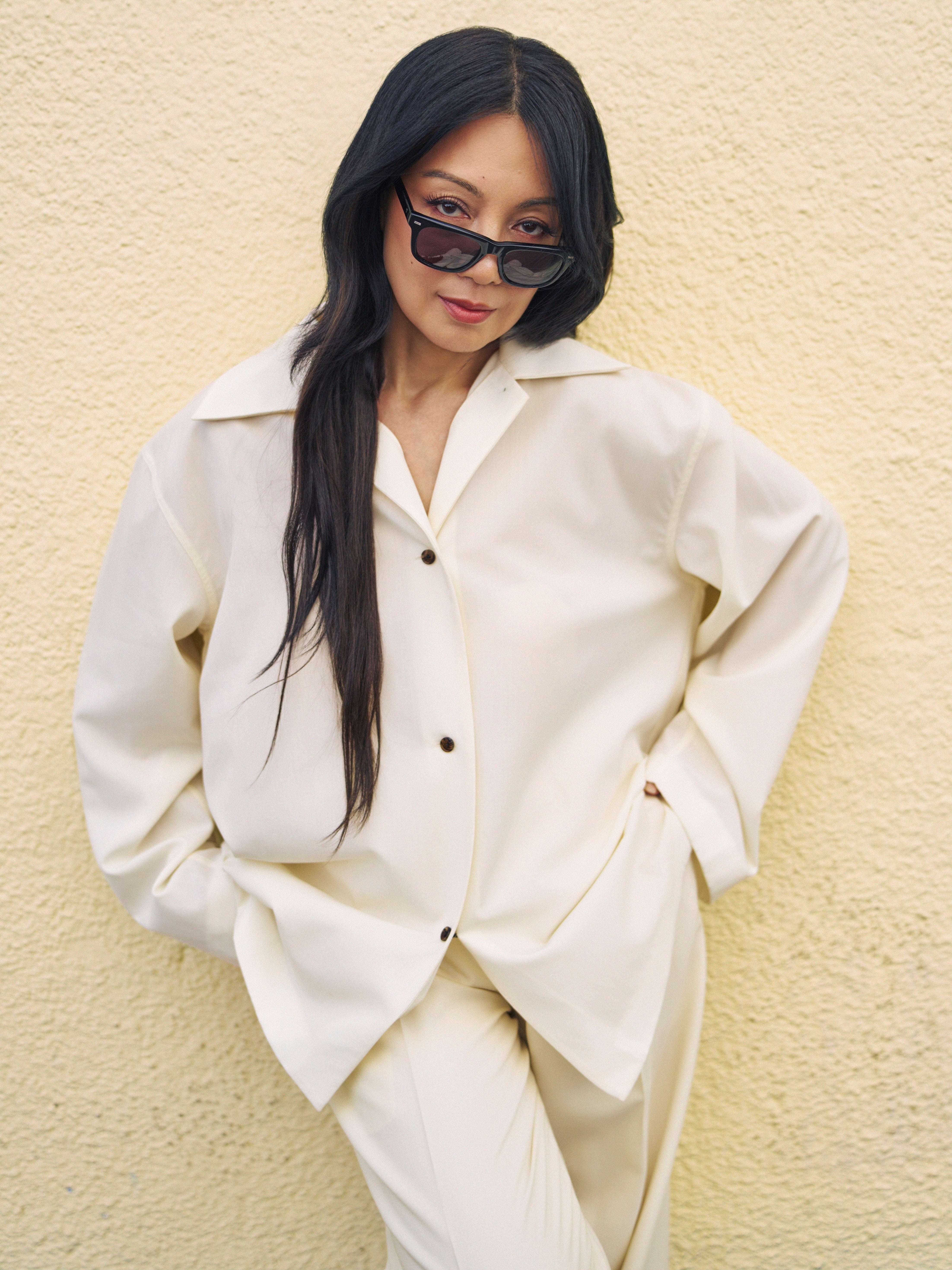 Person wearing a stylish oversized suit and cat-eye sunglasses poses confidently against a neutral background