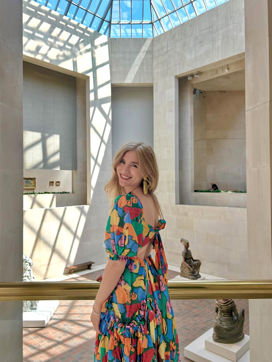 Woman in a vibrant, patterned dress smiles in a sunlit atrium with sculptures, suitable for stylish, artistic fashion pieces
