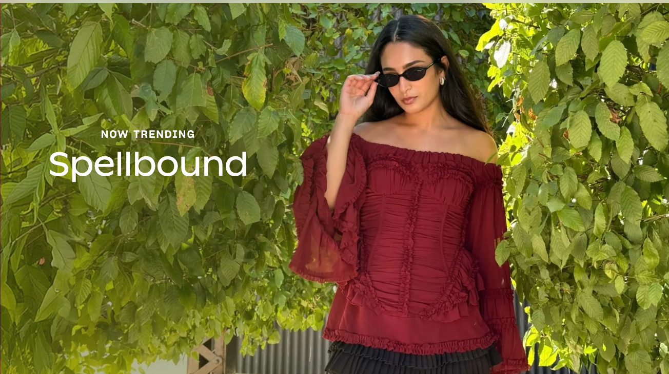 Woman in off-the-shoulder top with ruffled sleeves holds sunglasses, standing among greenery. Text: "Now Trending: Spellbound."