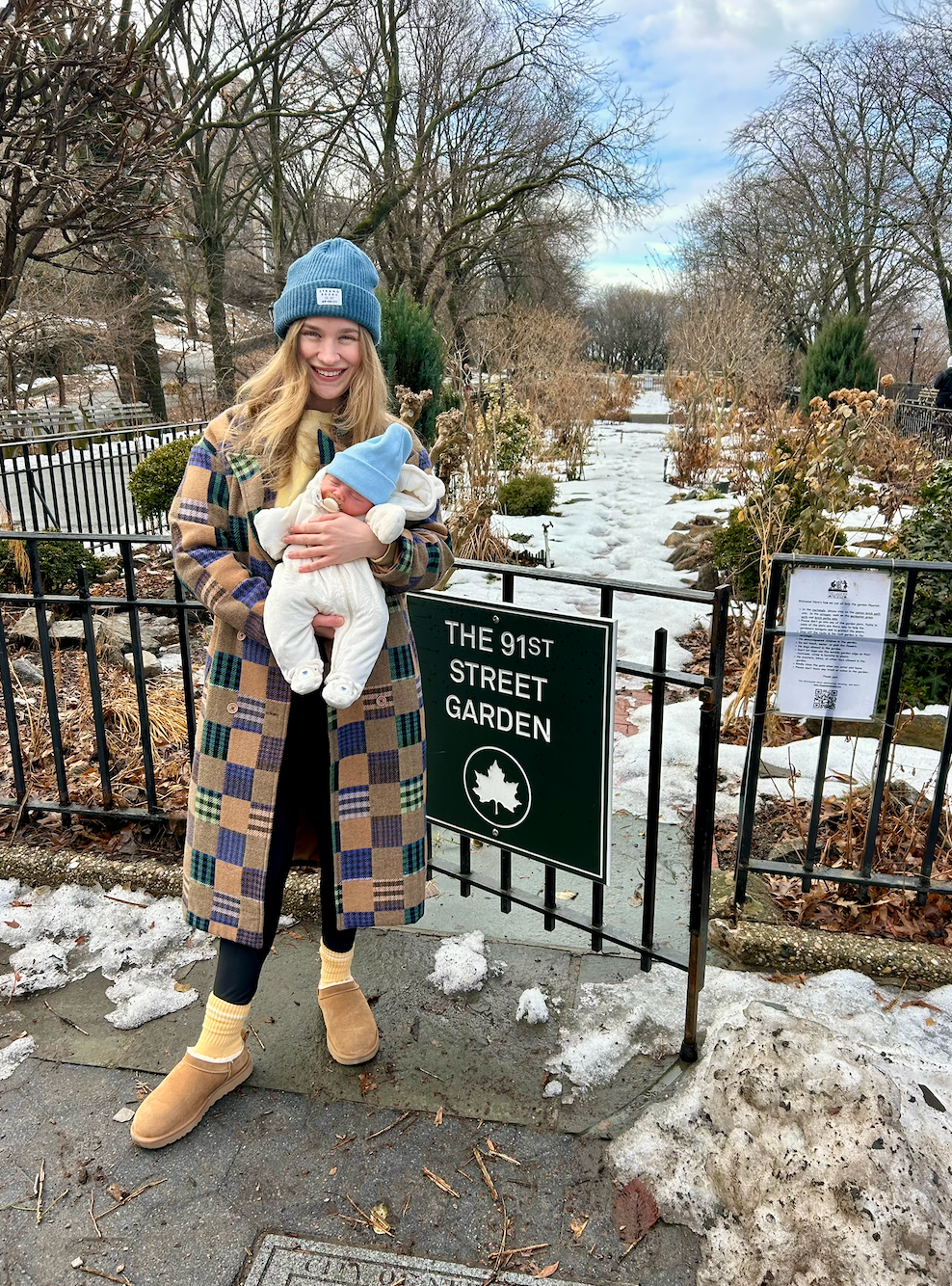 Person outdoors in a plaid coat and beanie holding a baby in a park near a sign reading "The 91st Street Garden."