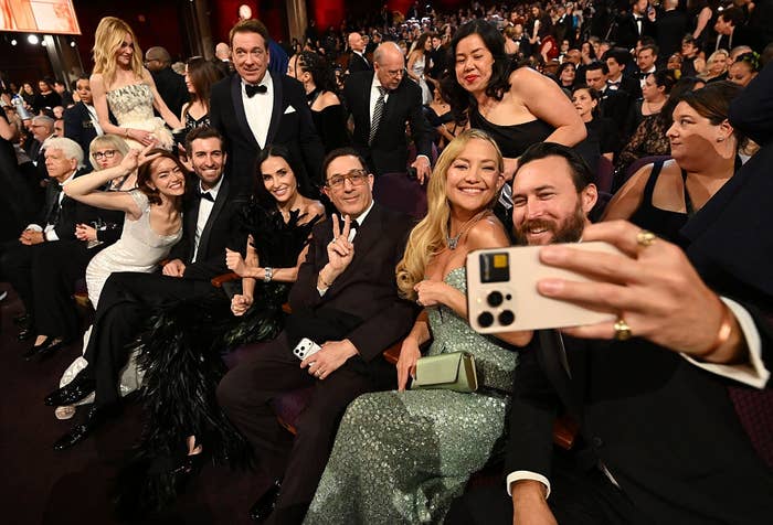 Group of celebrities in formal attire posing for a selfie at an event, surrounded by a seated audience
