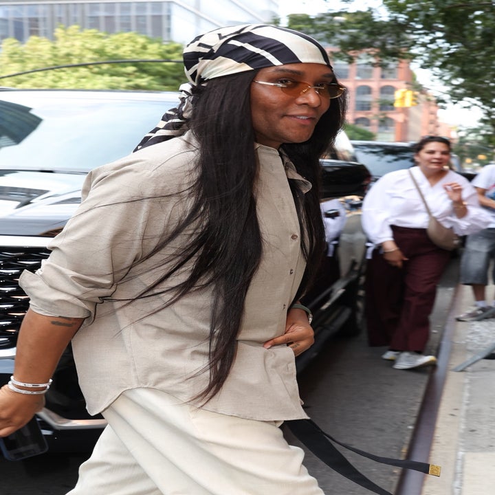 Person walking on a city street wearing a flowing outfit and headscarf, smiling towards onlookers. People in background behind a barrier