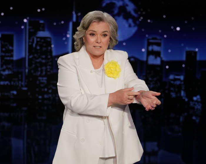 Person in a stylish white suit with a yellow flower brooch, gesturing on a nighttime TV show set with a city skyline backdrop
