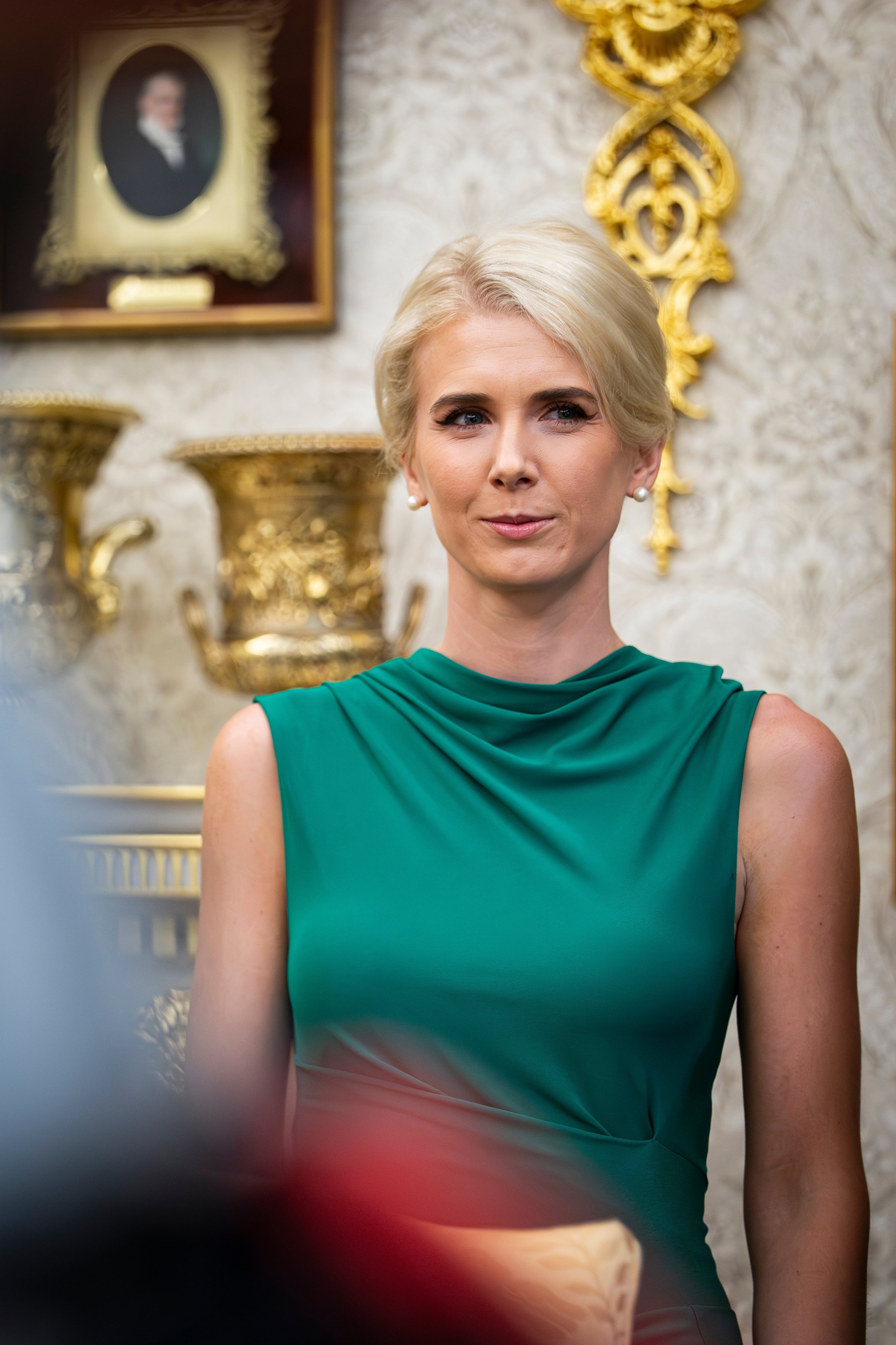 Person i elegant grønn kjole som står innendørs, med utsmykket dekor og innrammet portrett i bakgrunnen