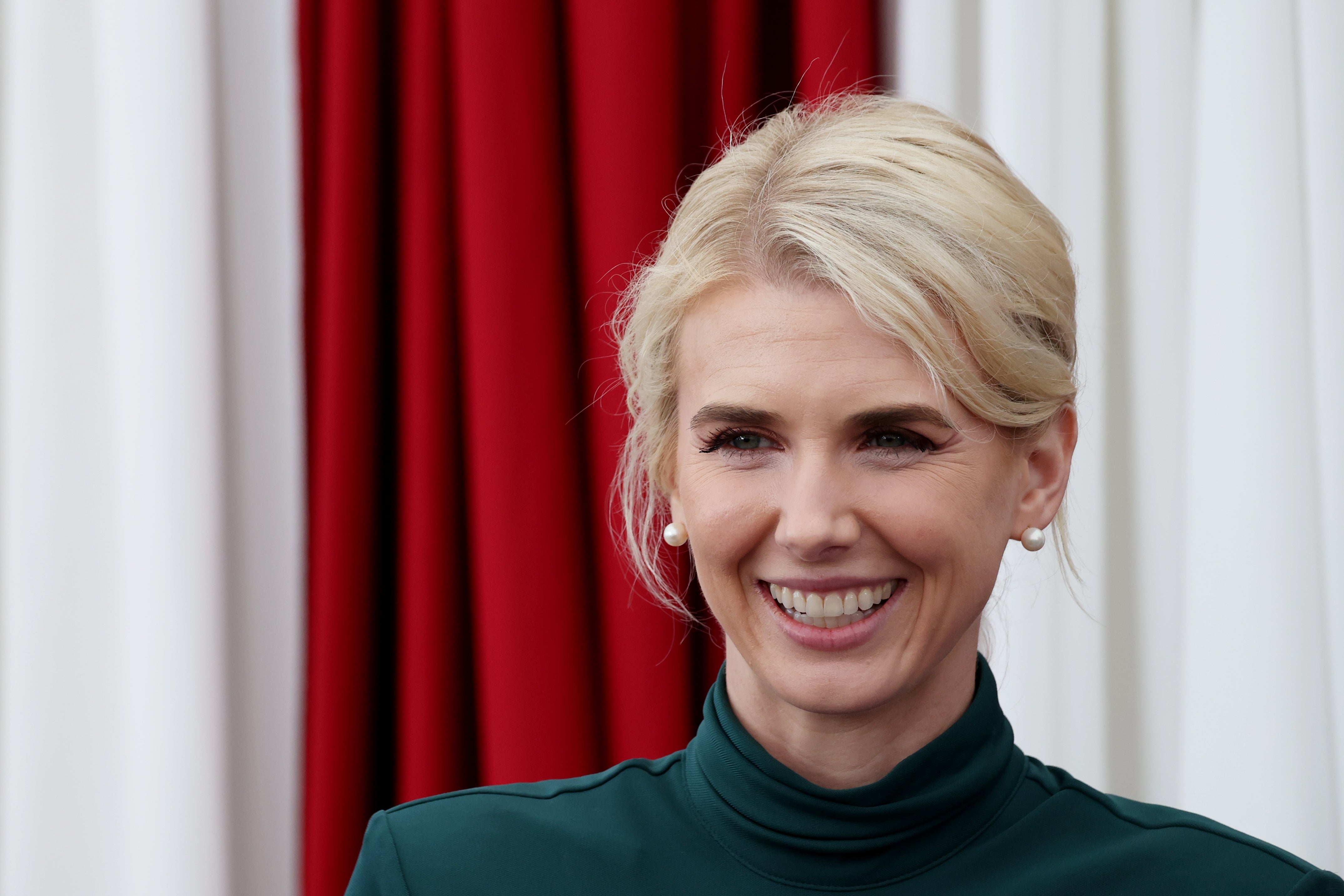 En person som smiler foran en bakgrunn med drapert rødt og hvitt stoff. De har kort blondt hår og har på seg et antrekk med høy hals