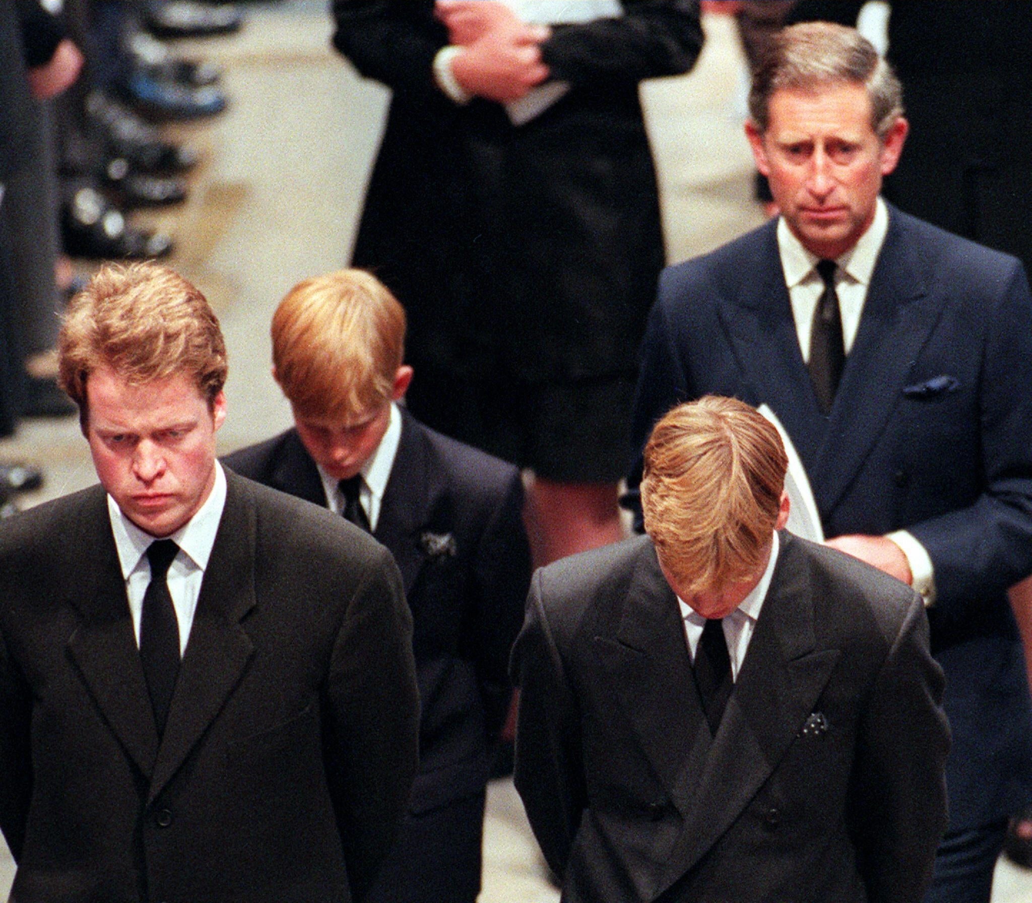 Four men in formal black suits and ties, standing solemnly with heads bowed during a somber event