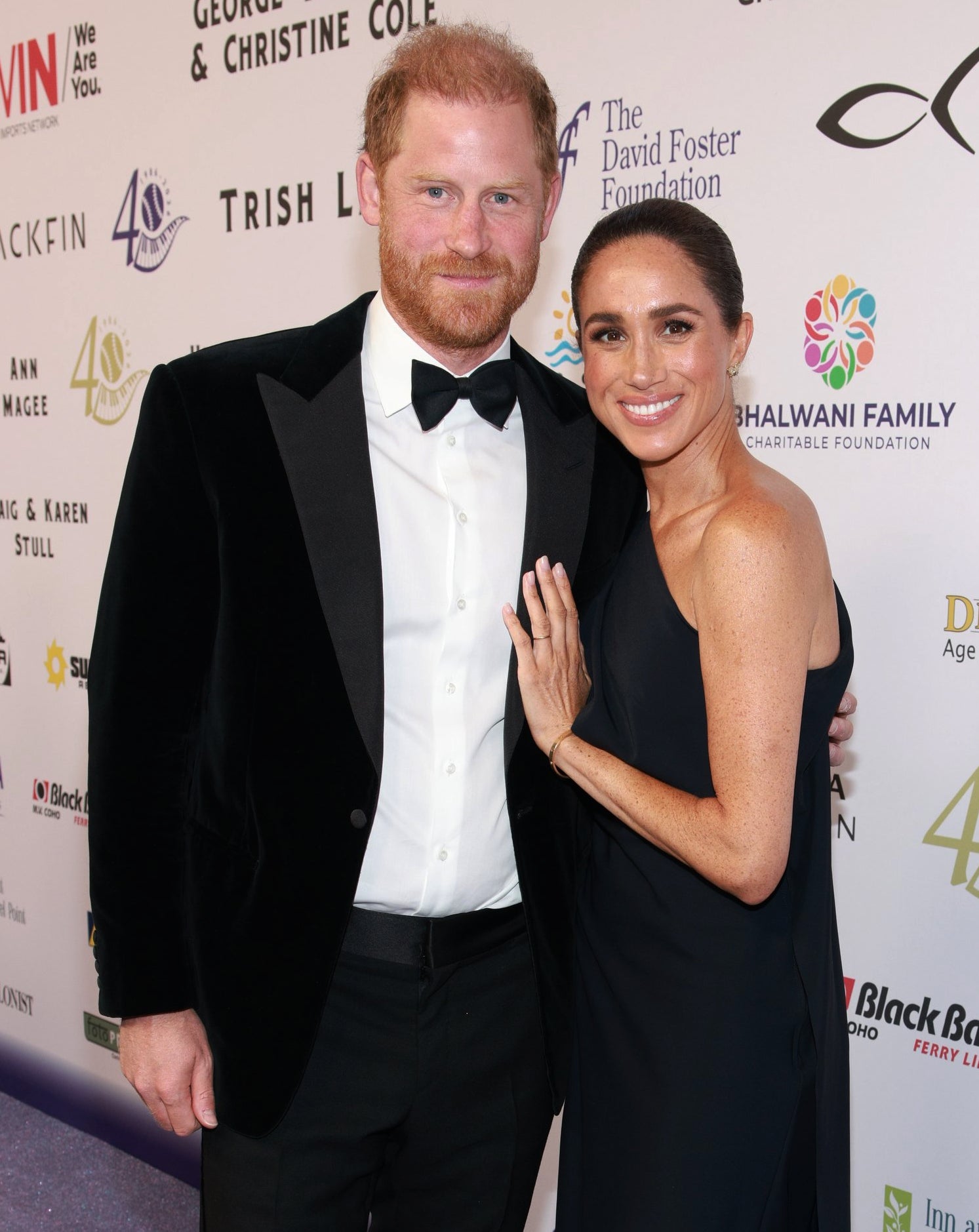 A man and a woman pose on a red carpet. The man is in a black tuxedo, and the woman is in a sleeveless black gown. Logos are visible in the background