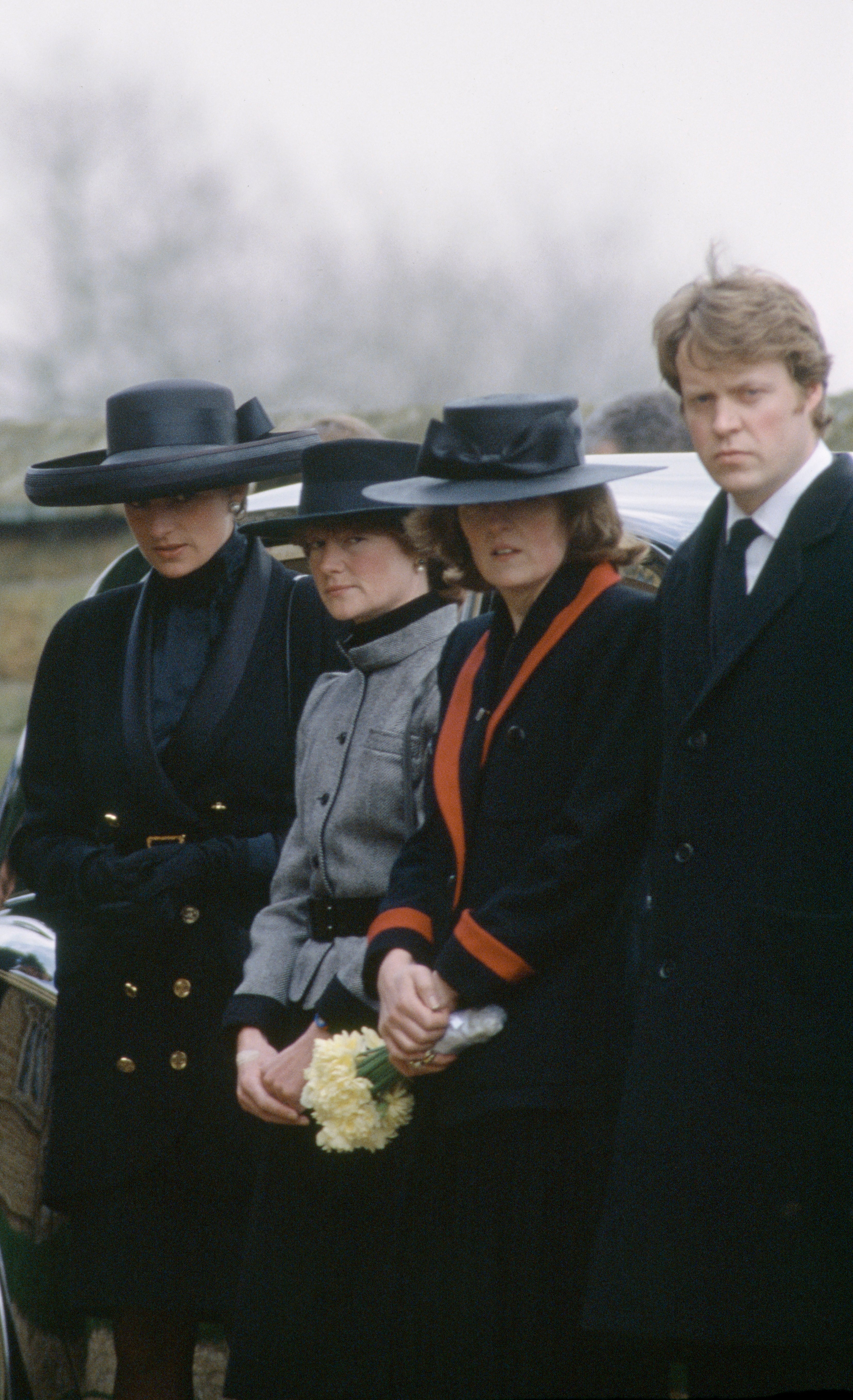 Four people at a formal outdoor event appear somber. Three women wear hats and coats, one holds flowers. A man is in a dark suit