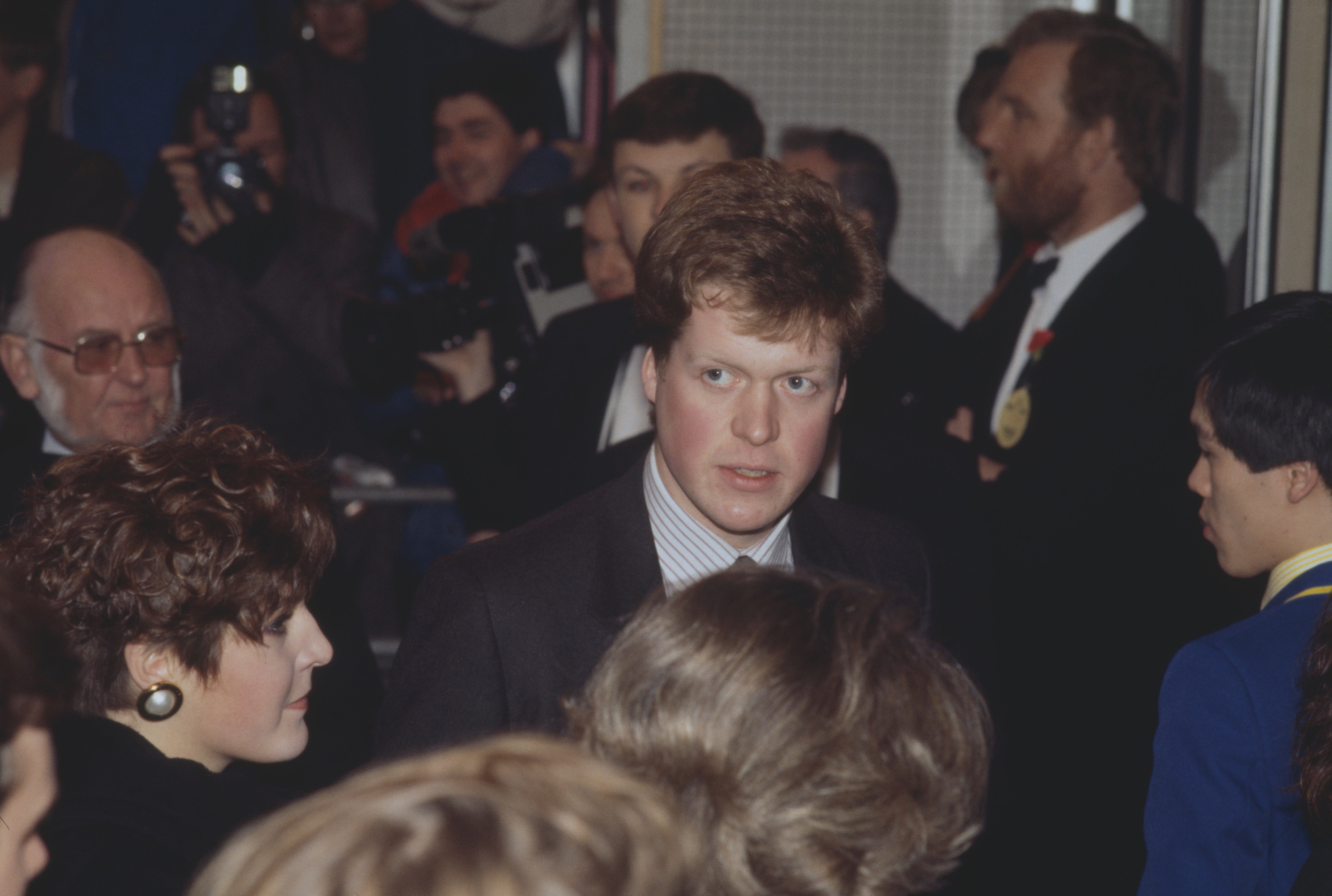A man in a suit at a formal event with people around, some taking photos