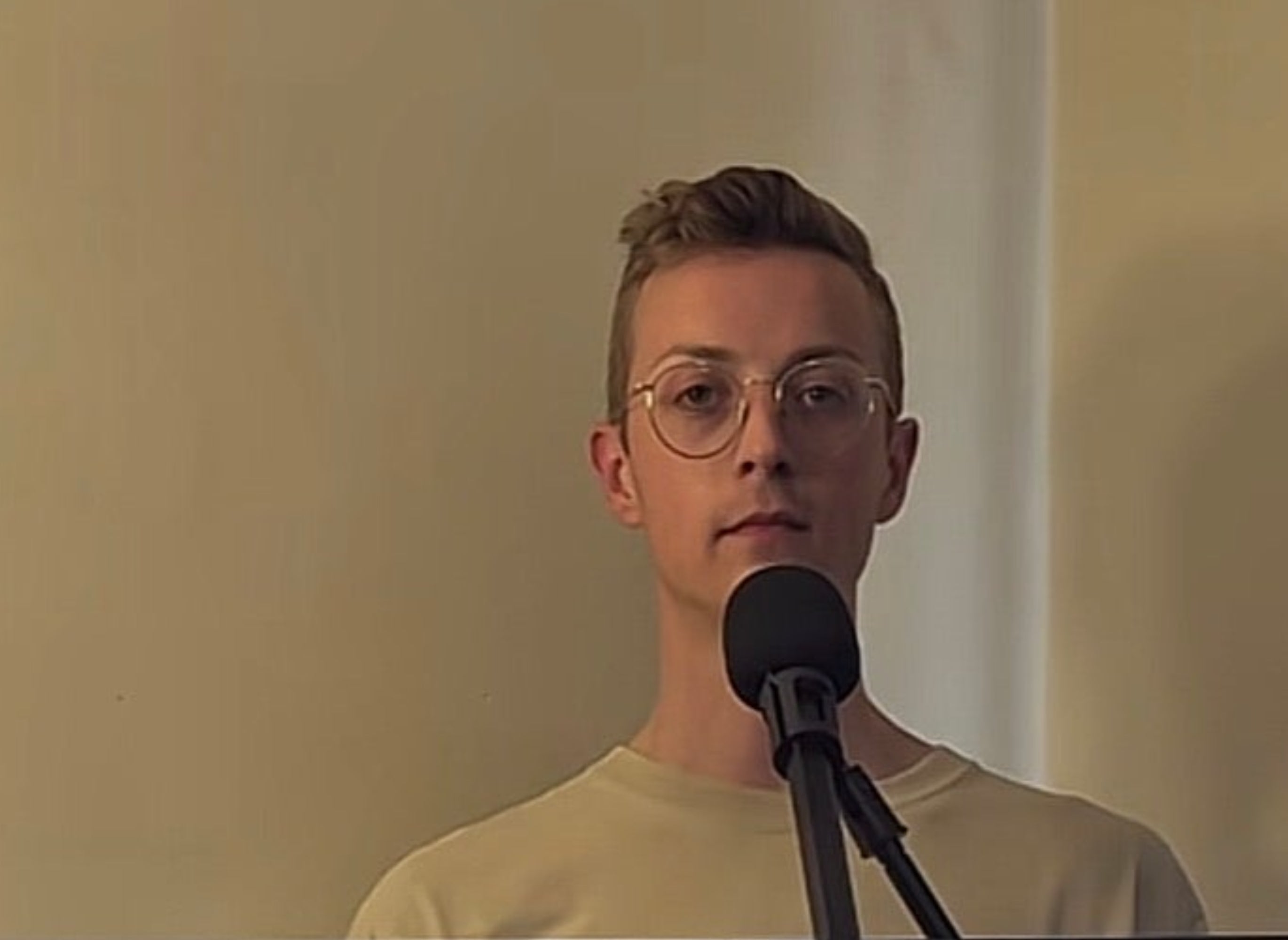Person som står innendørs ved en mikrofon, har på seg briller og en vanlig skjorte, ser ut til å snakke eller synge