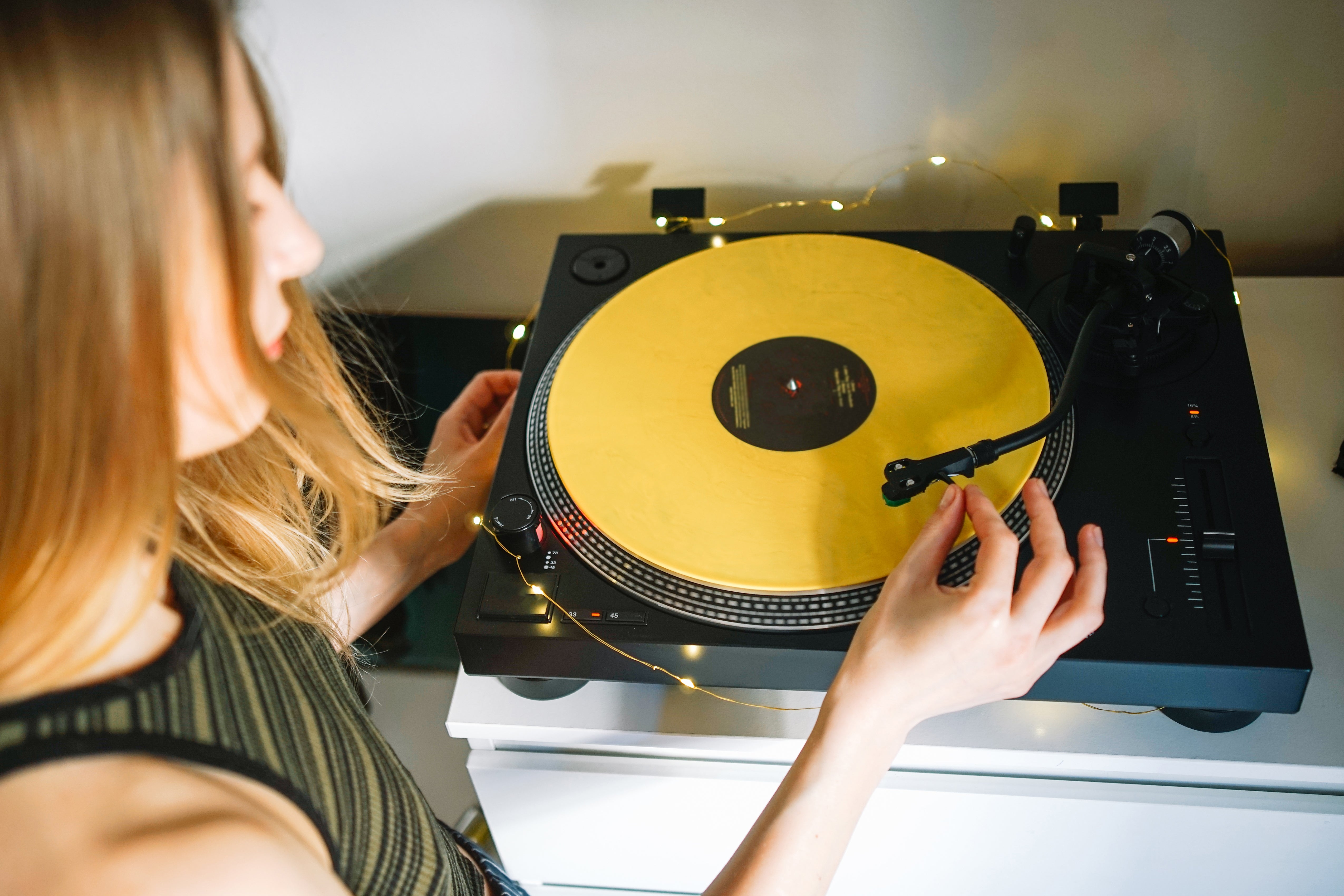 Person adjusting a turntable playing a yellow vinyl record, with warm lights around it. The scene suggests a casual, cozy atmosphere