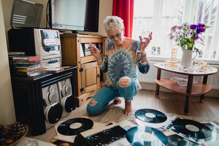 Person kneeling amid vinyl records, wearing a patterned blouse and ripped jeans, holding a drink, with a stereo and flowers nearby