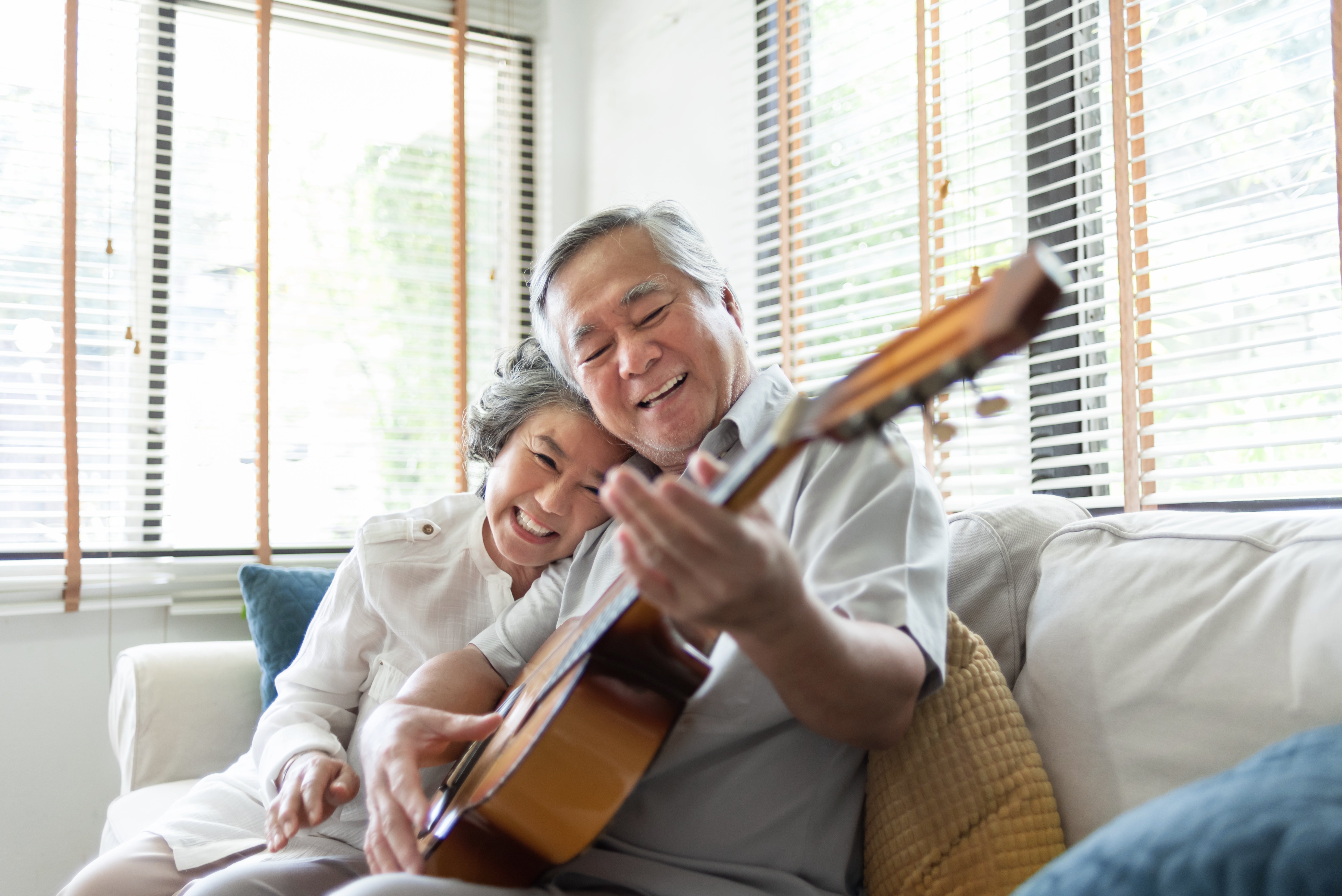 Older couple smiling and sitting together on a couch as one plays the guitar. Bright natural light filters in through the window blinds