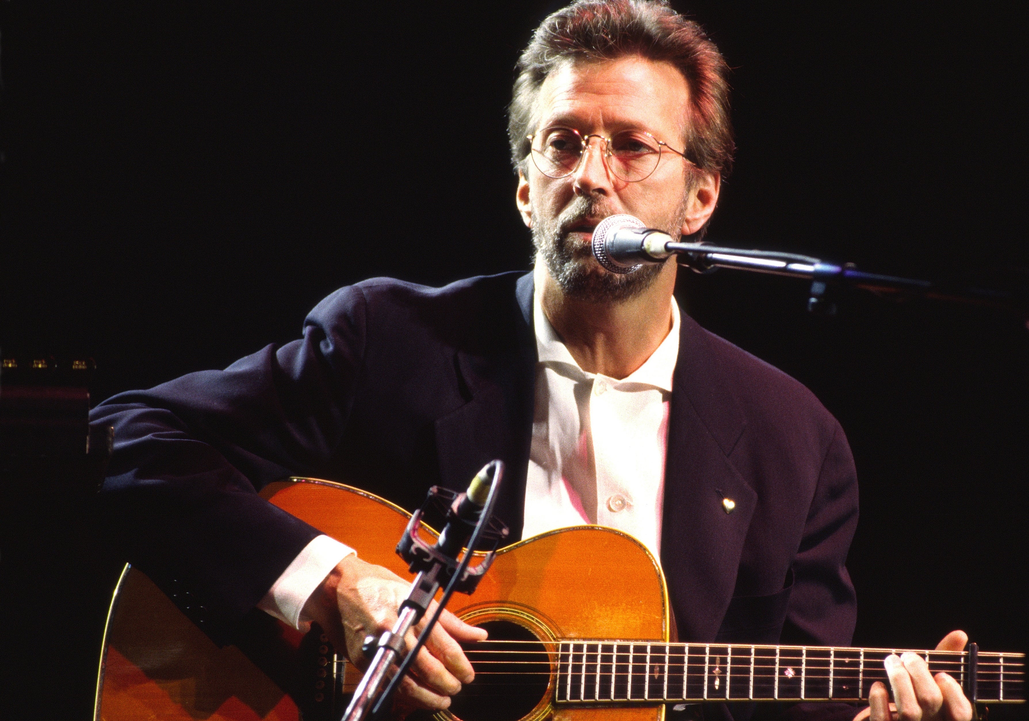 Musician playing an acoustic guitar, singing into a microphone, wearing a suit with an open-collared shirt