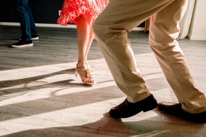 People dancing, focus on their feet and legs. One person wears a flowy skirt, the other wears slacks and dress shoes, both move energetically