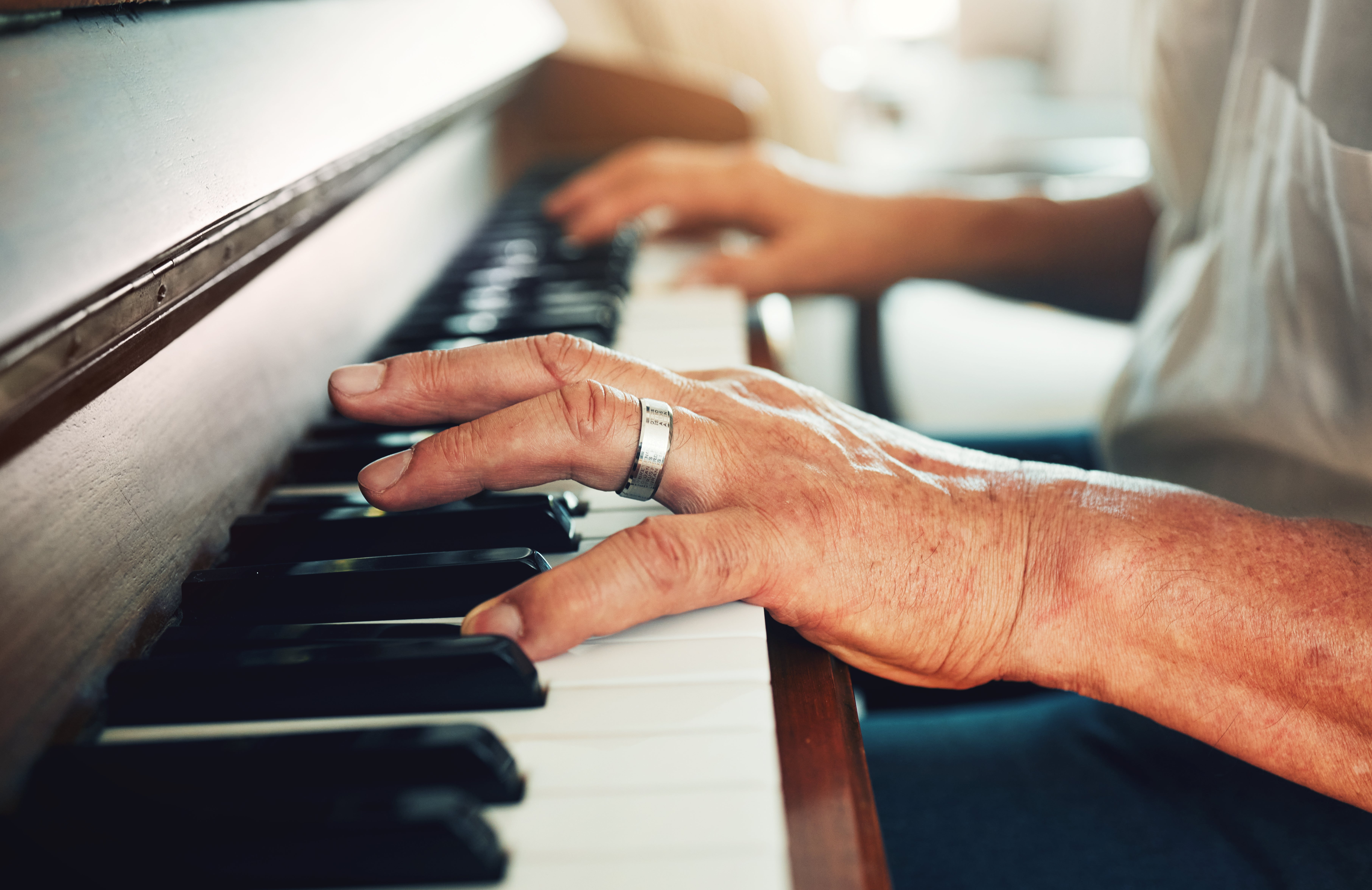 A person wearing a silver ring plays a piano, focusing on the keys, with sunlight illuminating their hands