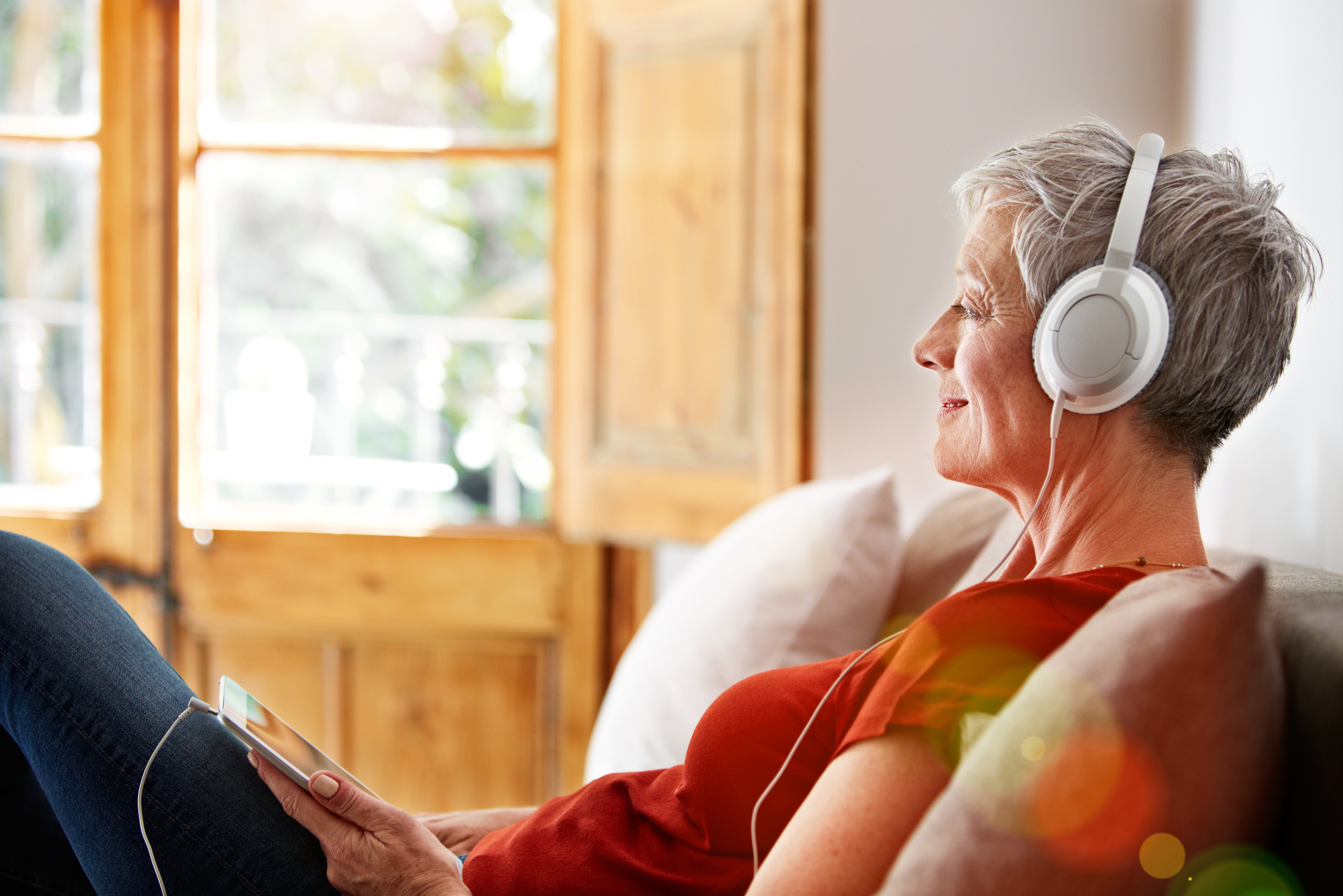 Elderly woman relaxing on a couch, listening to headphones, holding a tablet, looking content
