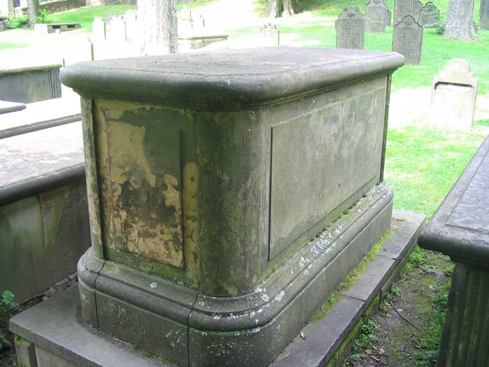 Weathered, moss-covered stone tomb in a cemetery surrounded by gravestones and grass