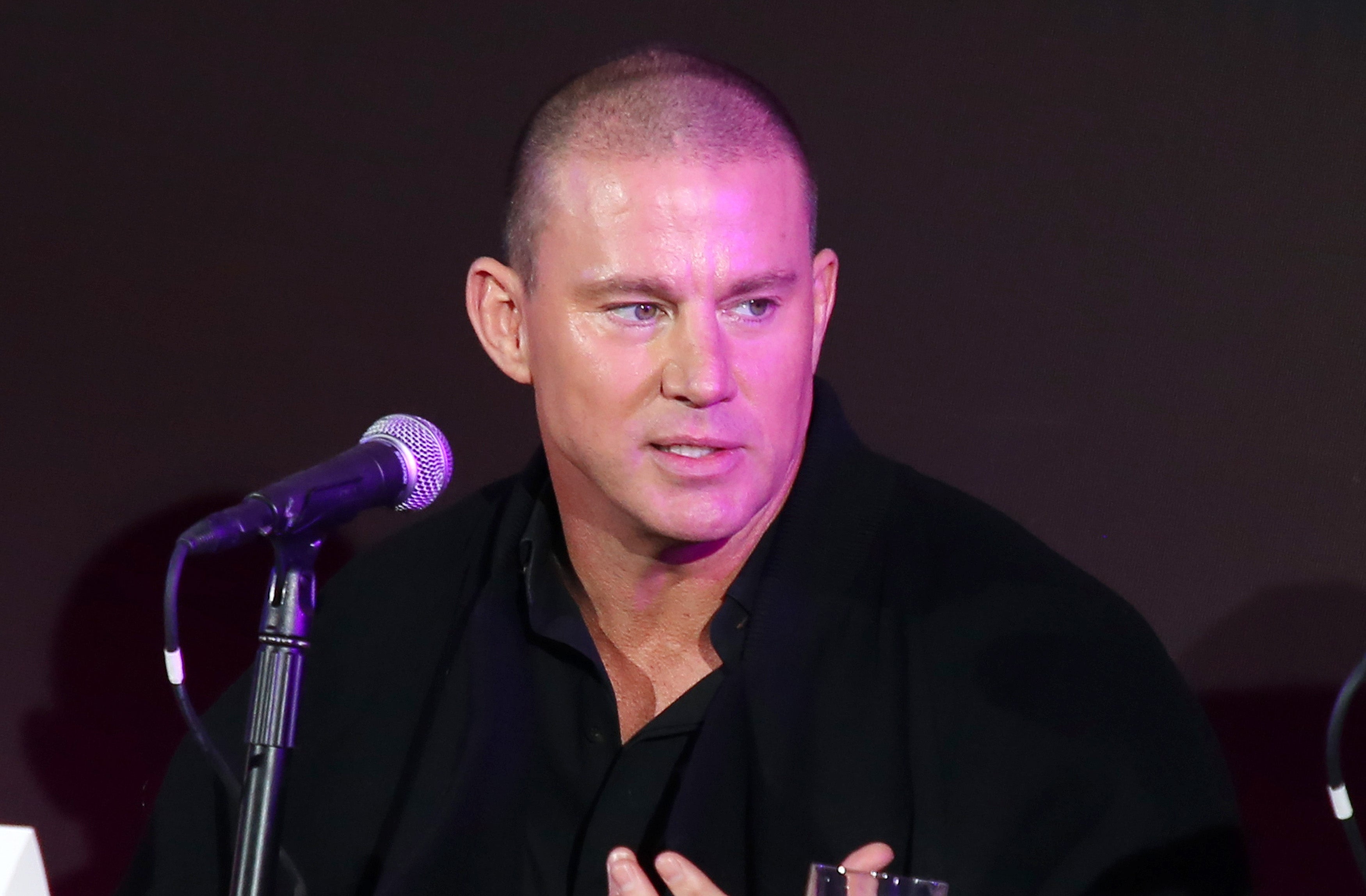 A person speaking into a microphone at an event, wearing a black outfit with a casual, confident style