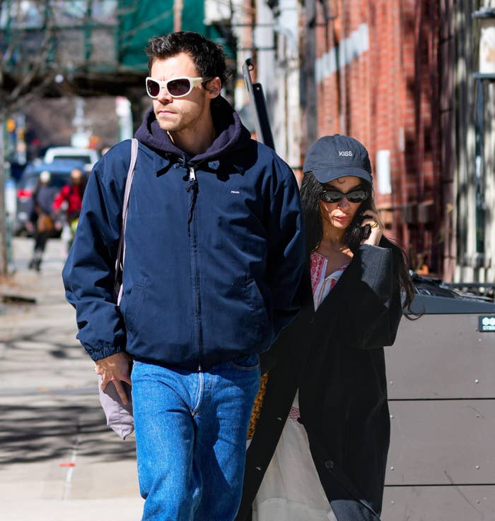 Two people walking outside, one in a navy jacket and sunglasses, the other in a black coat and cap. Casual urban setting