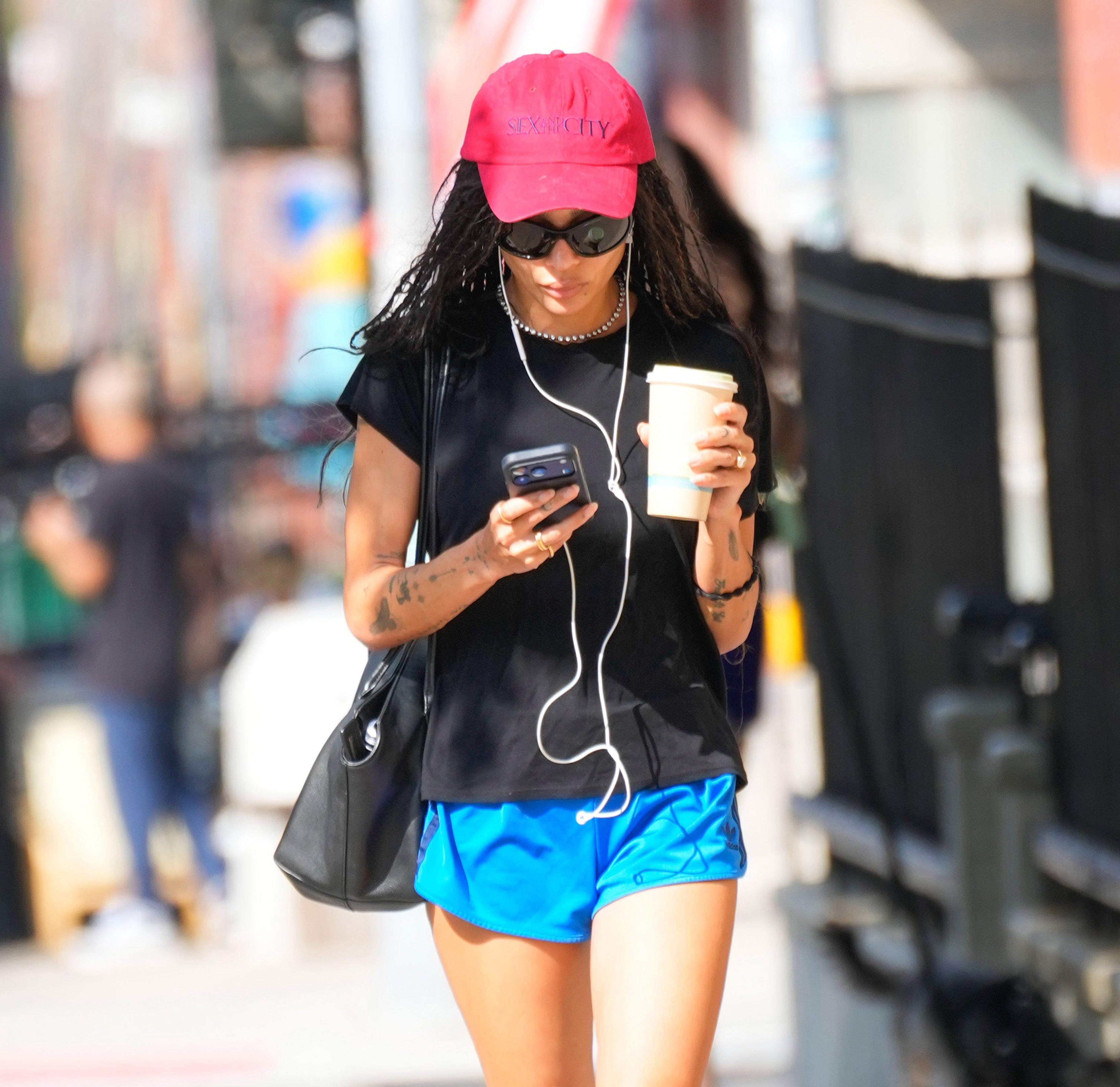 Person walking outside, wearing a casual outfit with a red cap, sunglasses, and earphones, holding a phone and a coffee cup